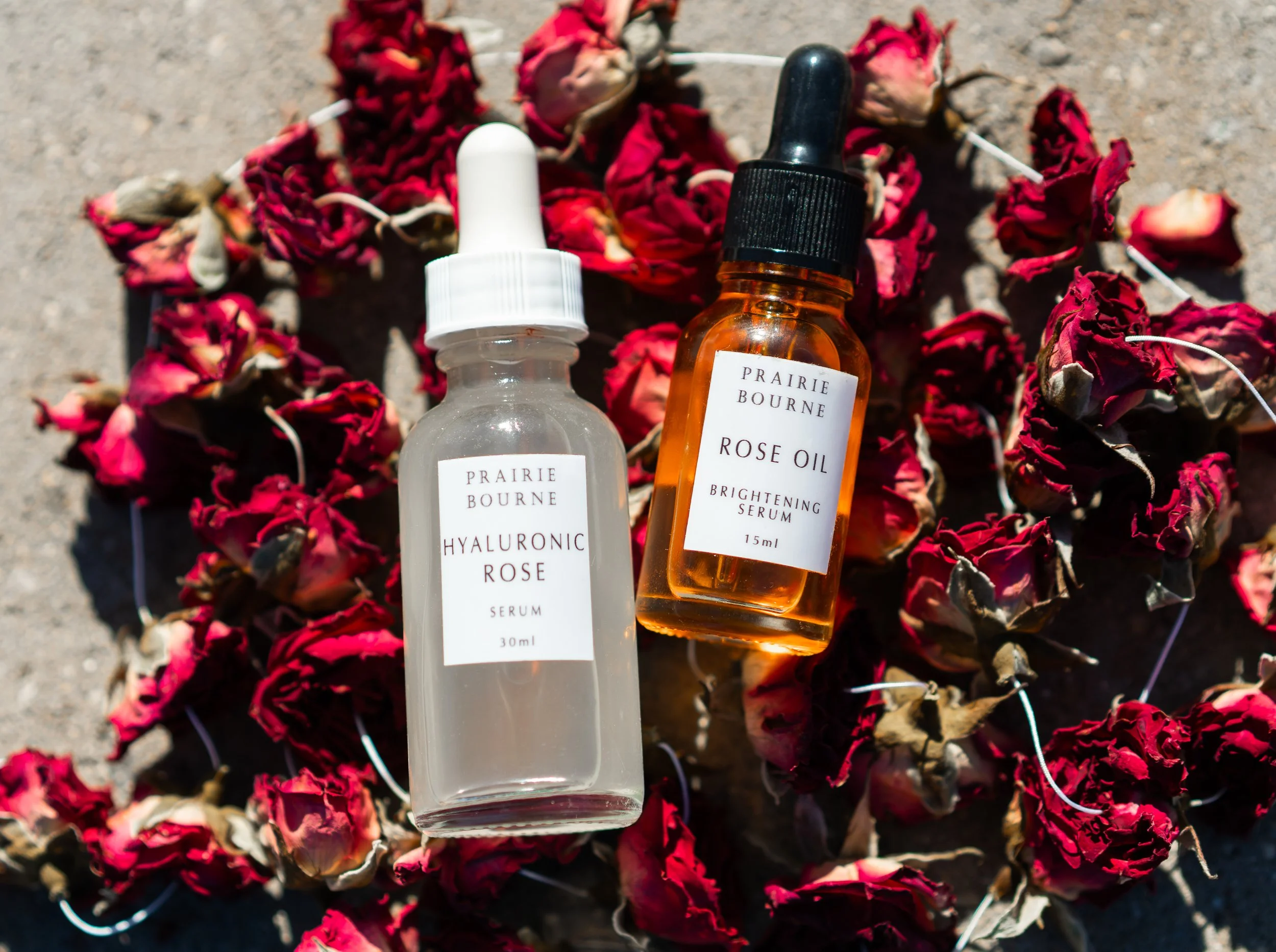 Two bottles of Prairie Bourne skincare products, one labeled Hyaluronic Rose Serum and the other Rose Oil Brightening Serum, resting on a surface surrounded by dried red rose petals.