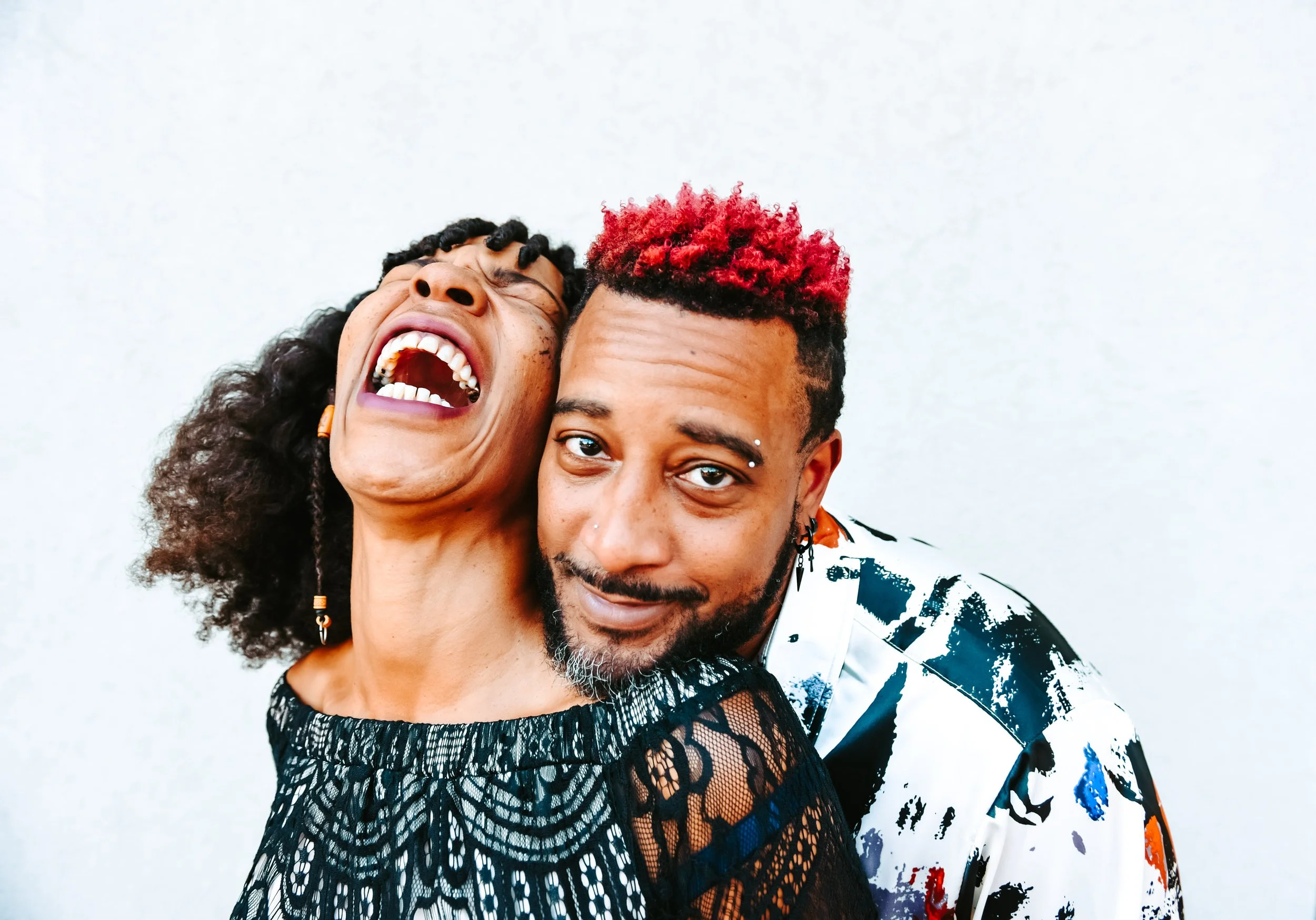 Two people close together, smiling and laughing, against a plain white background, one with curly hair and the other with dyed red hair.