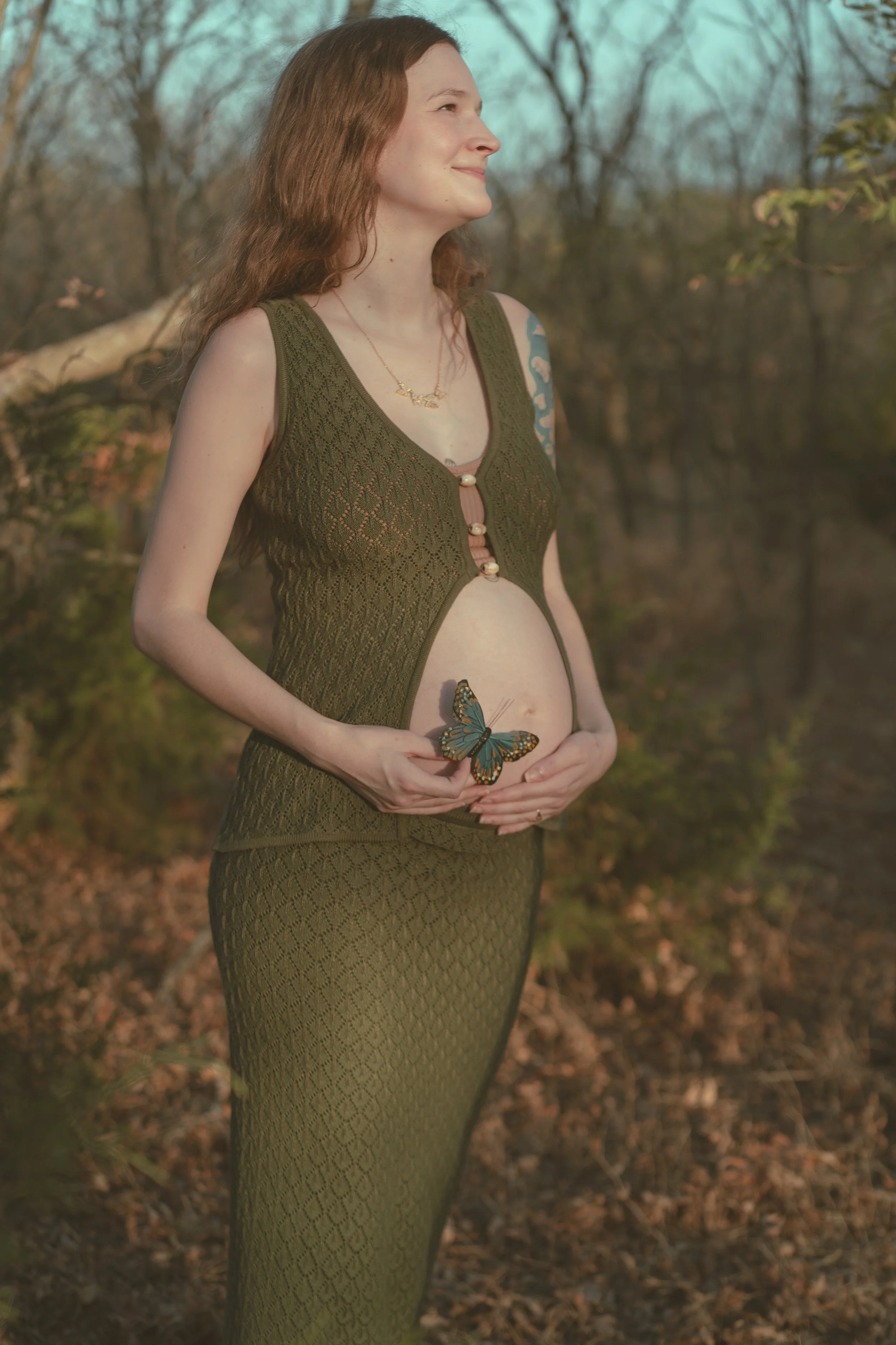 A pregnant woman with red hair standing outdoors during daytime, smiling and holding her belly with a butterfly perched on her hand, in a natural setting with trees and sunlight.