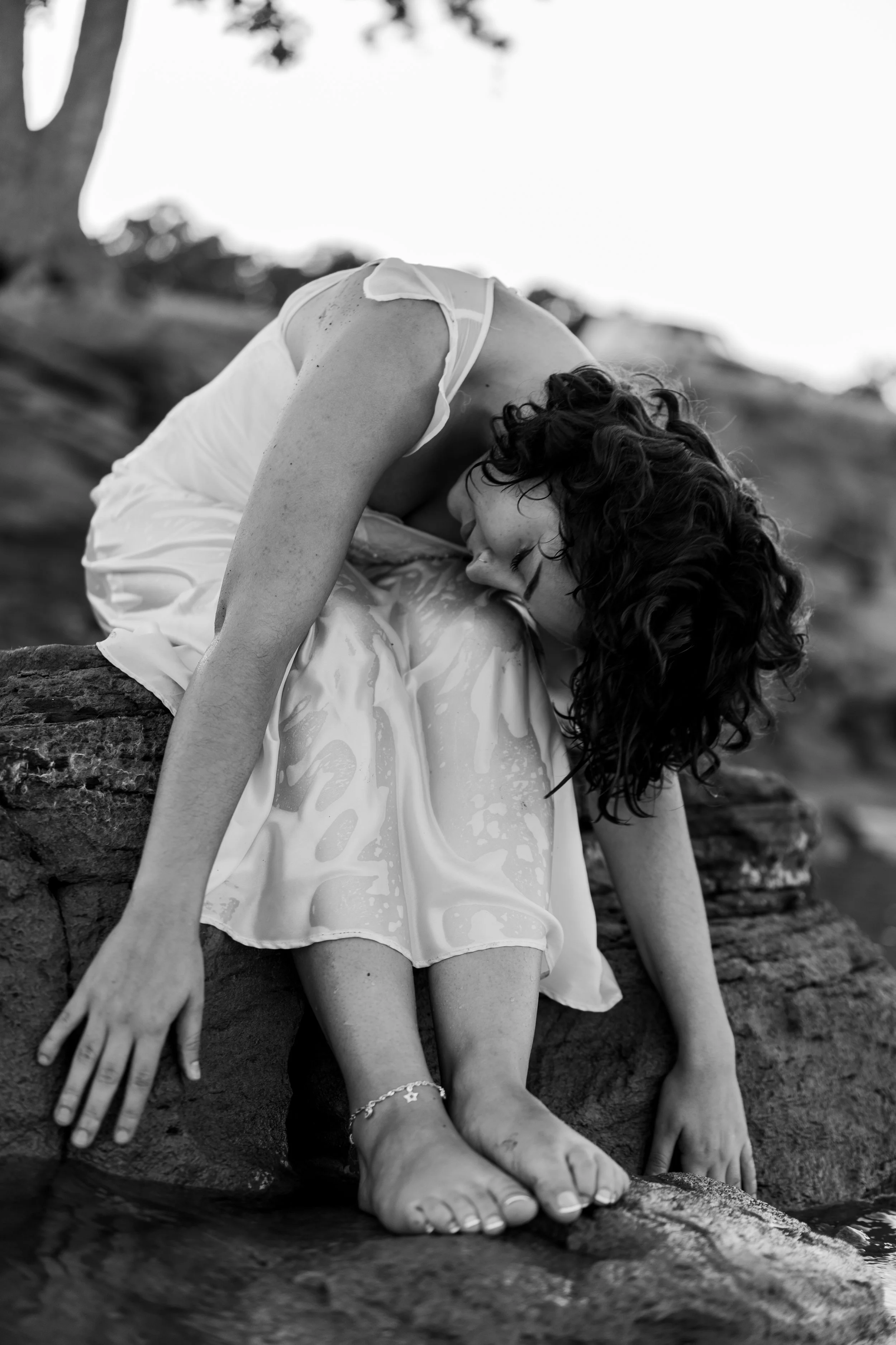 A black and white photo of a woman with curly hair in a wet dress sitting on rocks near water, with her head resting on her knees.