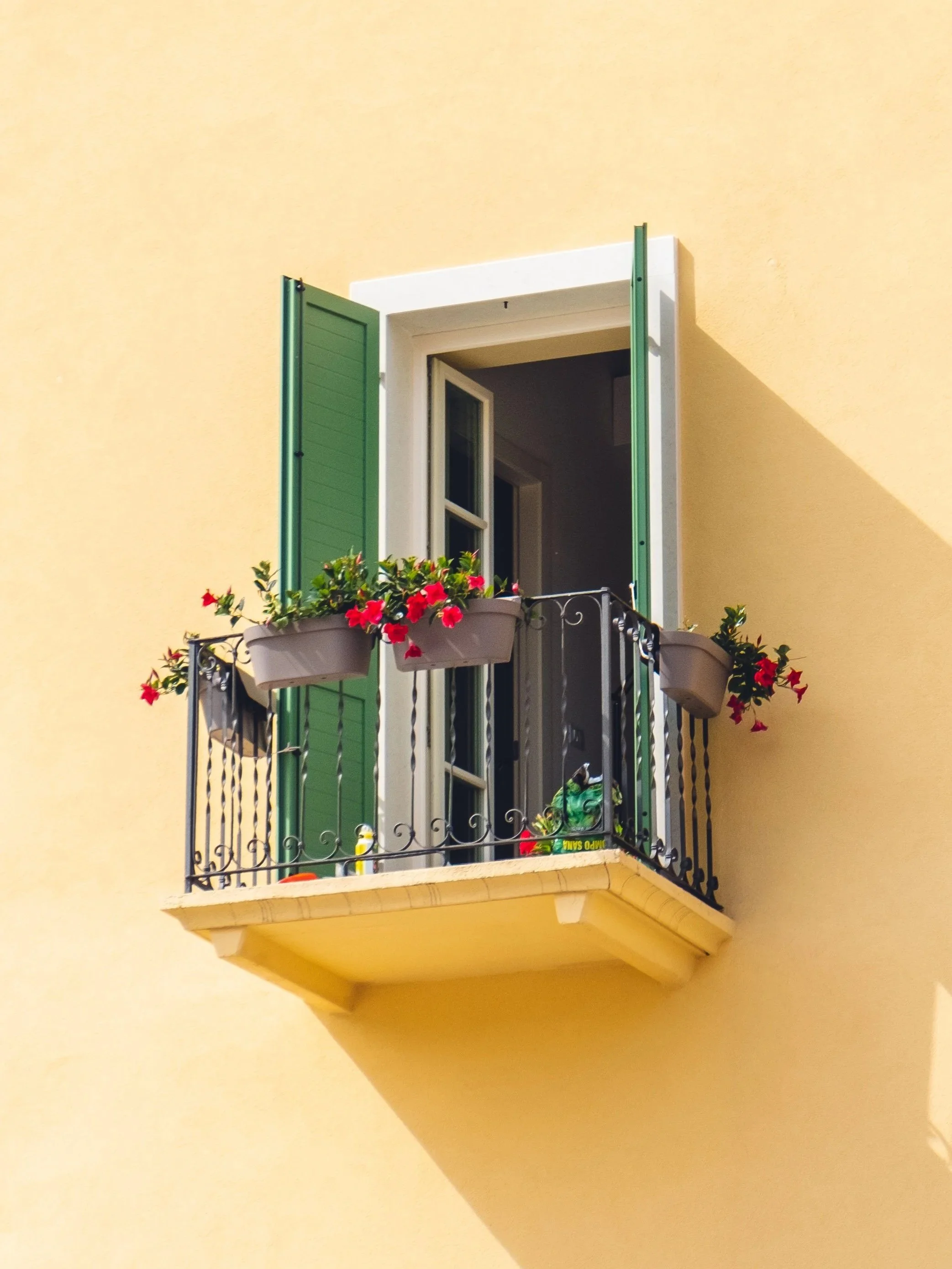 A small balcony with black iron railing, two flower pots with red flowers, green shutters, and an open window on a yellow building wall.