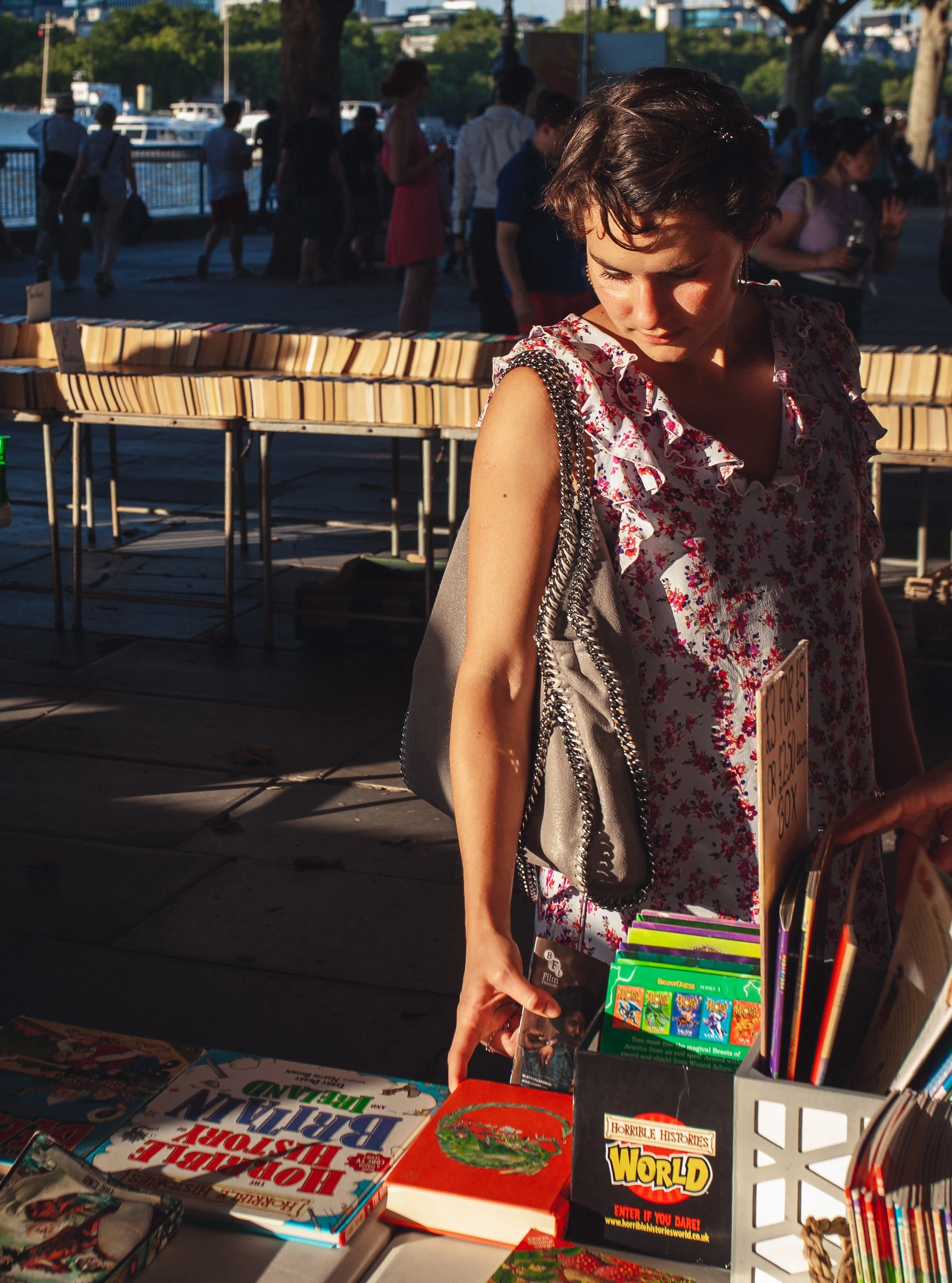 A woman shopping at an outdoor market during the daytime, looking at books displayed on a table. She has short dark hair, is wearing a sleeveless floral top, and carries a handbag with a chain strap. In the background, several people are walking near
