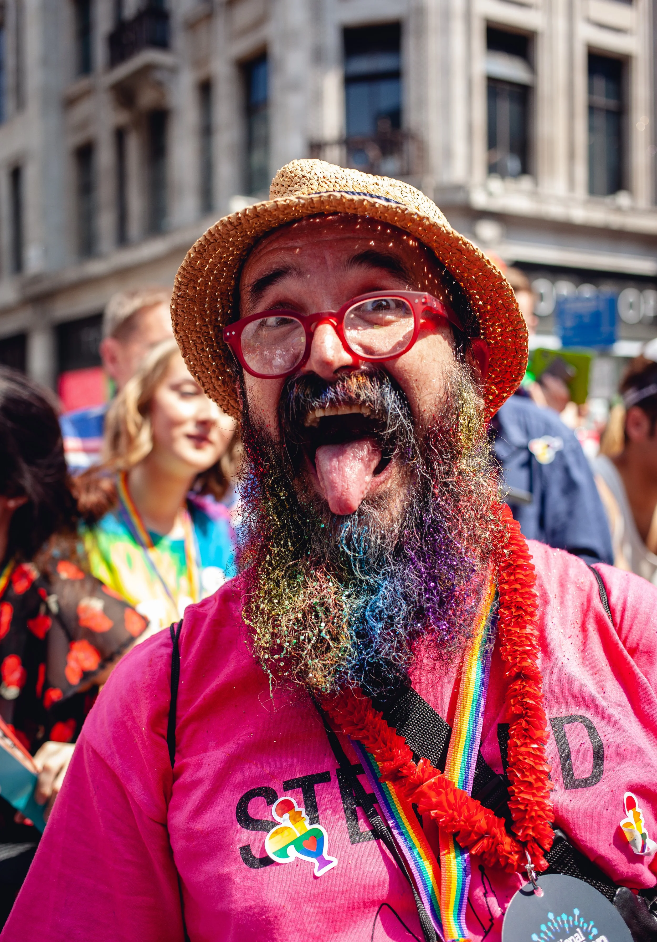 A man with a beard, wearing red glasses, a straw hat, and a pink t-shirt that says 'STAND', at a pride parade, sticking out his tongue and smiling, surrounded by people in colorful outfits.