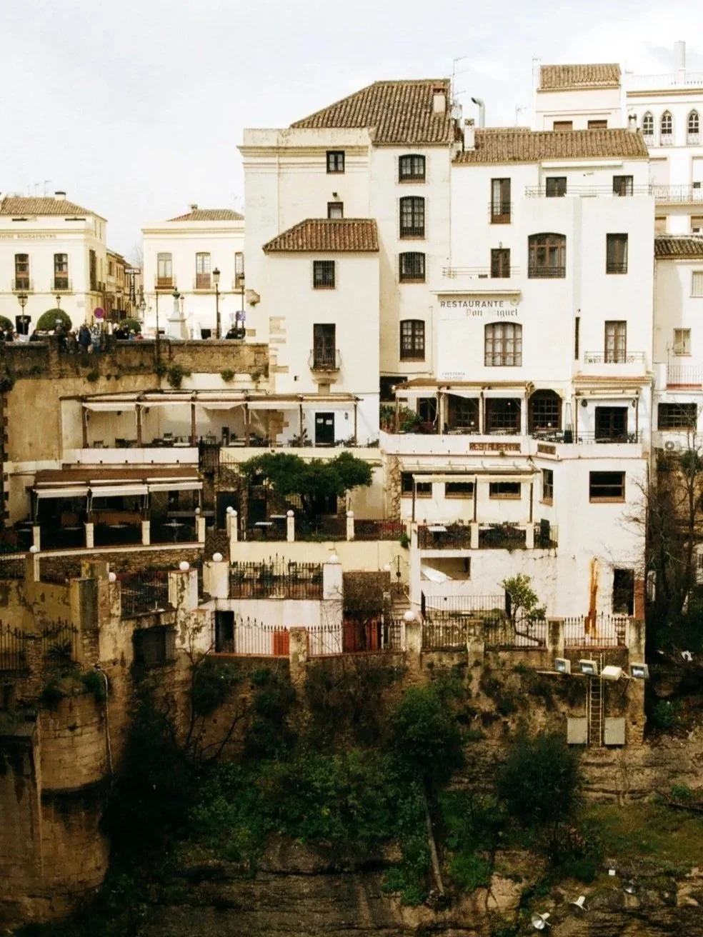 A hillside scene with white European-style buildings, some with balconies and red-tiled roofs, and a restaurant on the right side at the middle level. There are trees, stairs, and a small fenced area at the bottom.