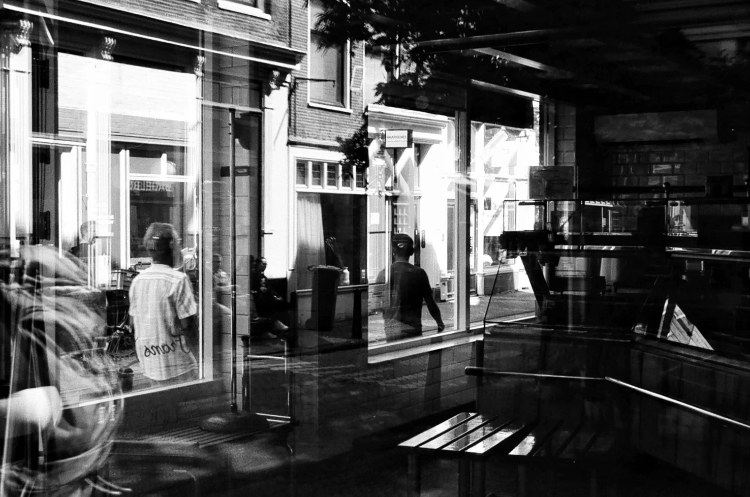 Reflection of people sitting and walking outside a shop window, with a woman in a striped shirt and a man in dark clothing visible among the reflections, and the street scene including buildings and trees.