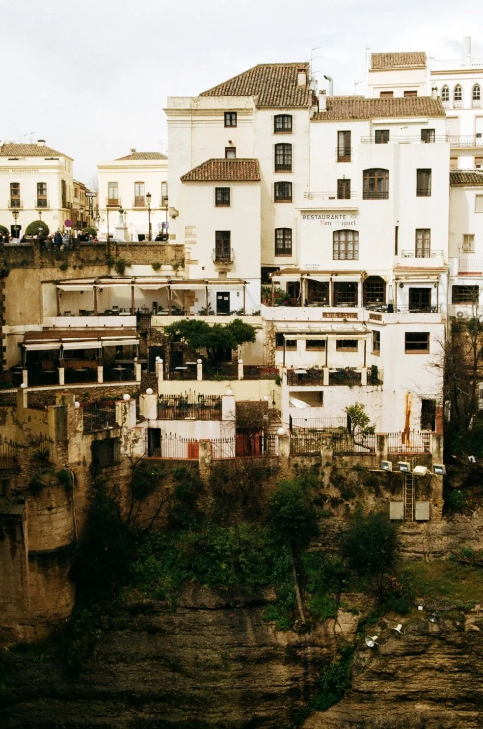 Multi-story white and beige buildings on a hillside with outdoor terraces and dining areas, overlooking a deep ravine with greenery.