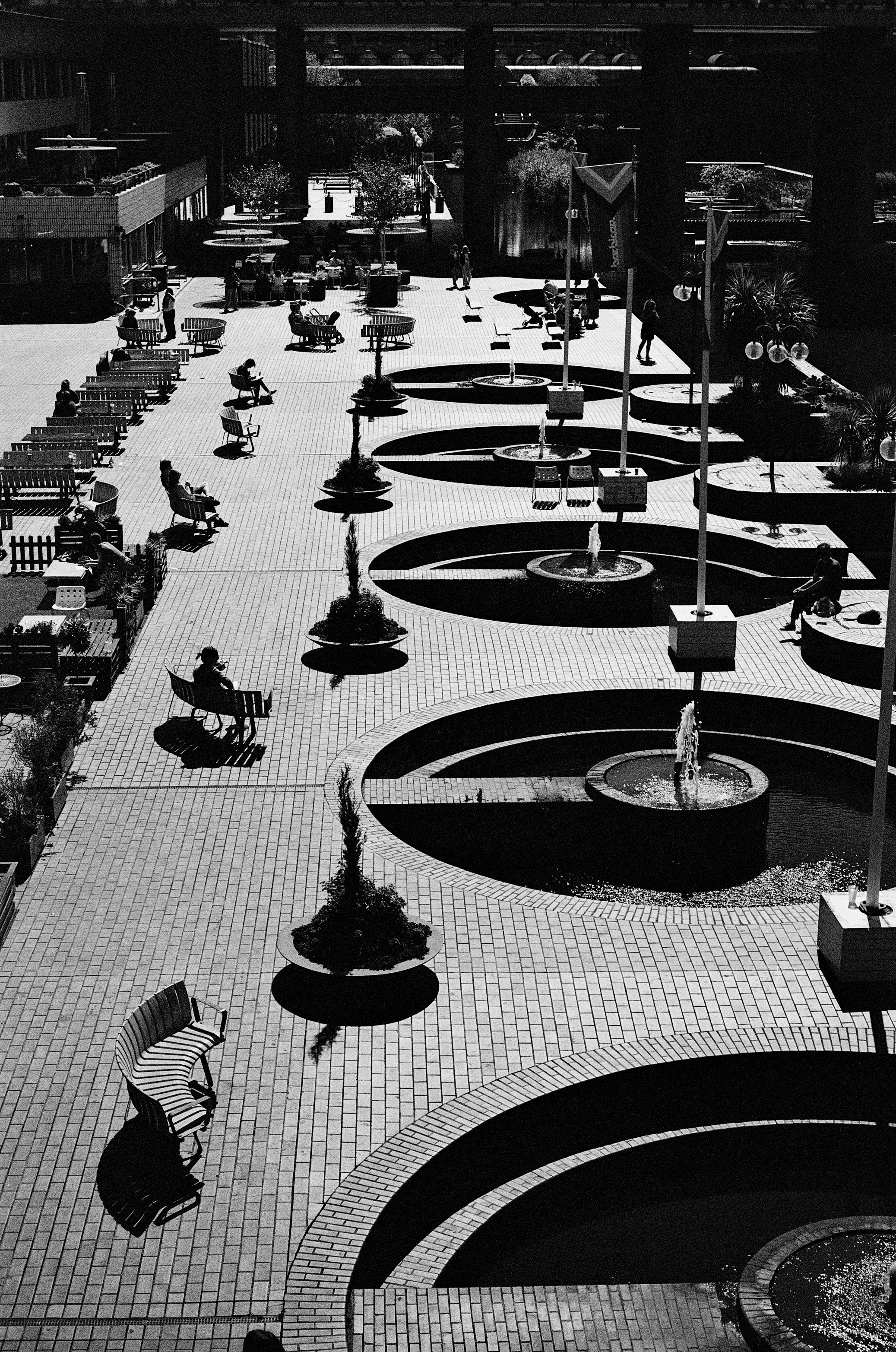 Black and white photo of an outdoor plaza with benches, trees, and fountains, and a few people walking or sitting.
