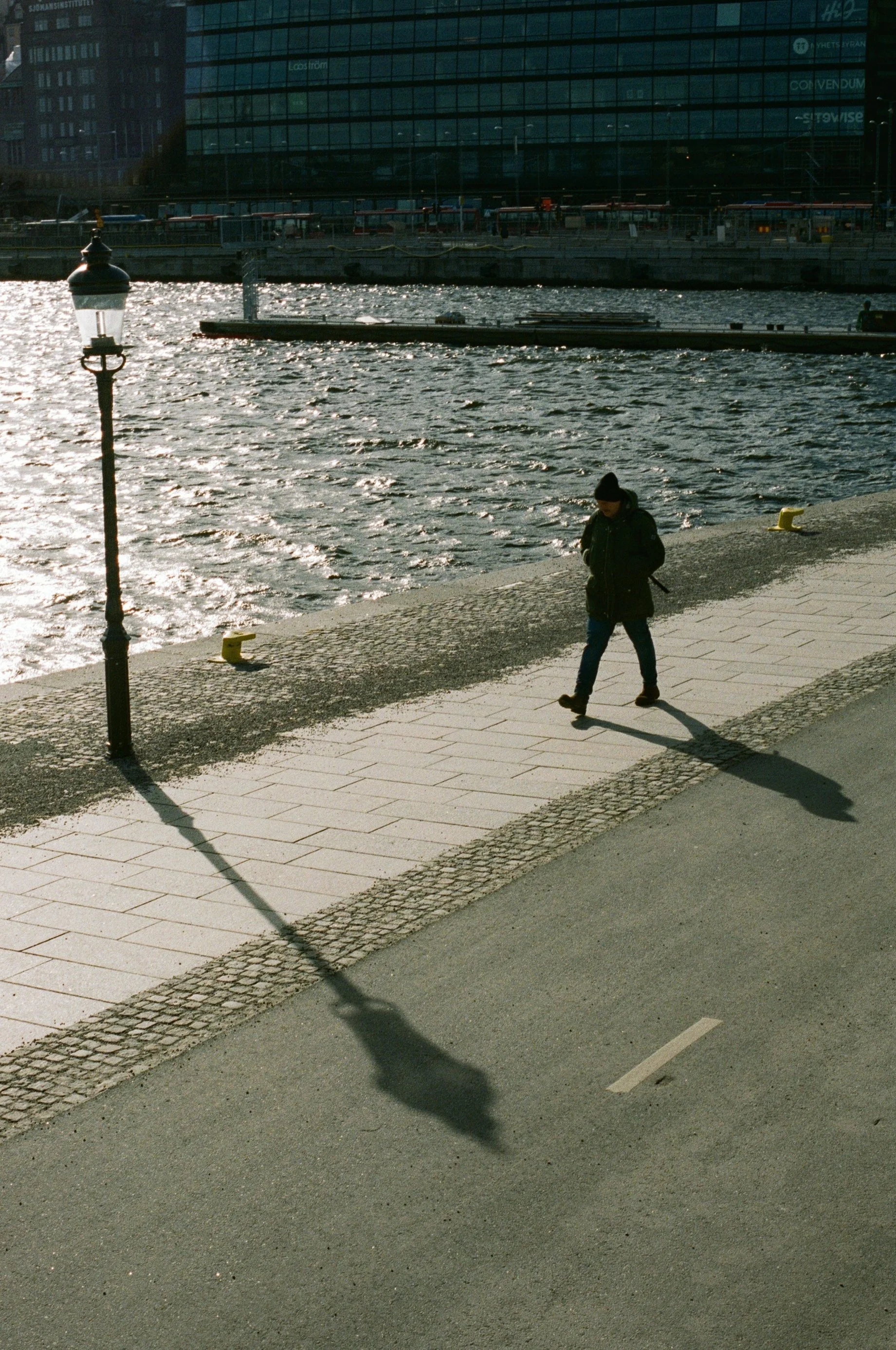 Person walking along a waterfront promenade on a sunny day, with a lamppost casting a long shadow and a large modern building across the water in the background.