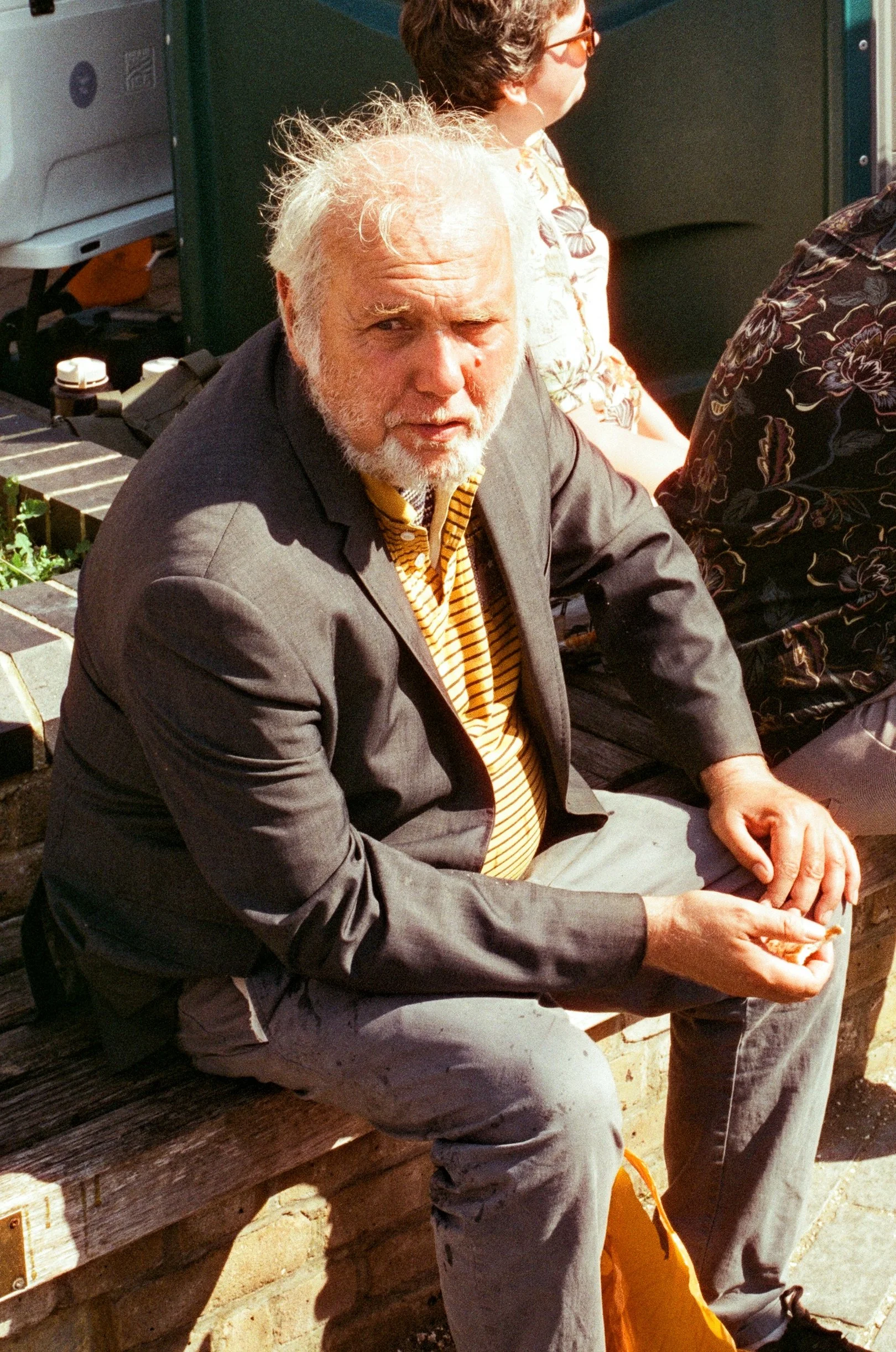 An elderly man with white hair and a beard, wearing a gray blazer and a yellow striped shirt, sitting on a wooden bench outdoors, with other people visible in the background.
