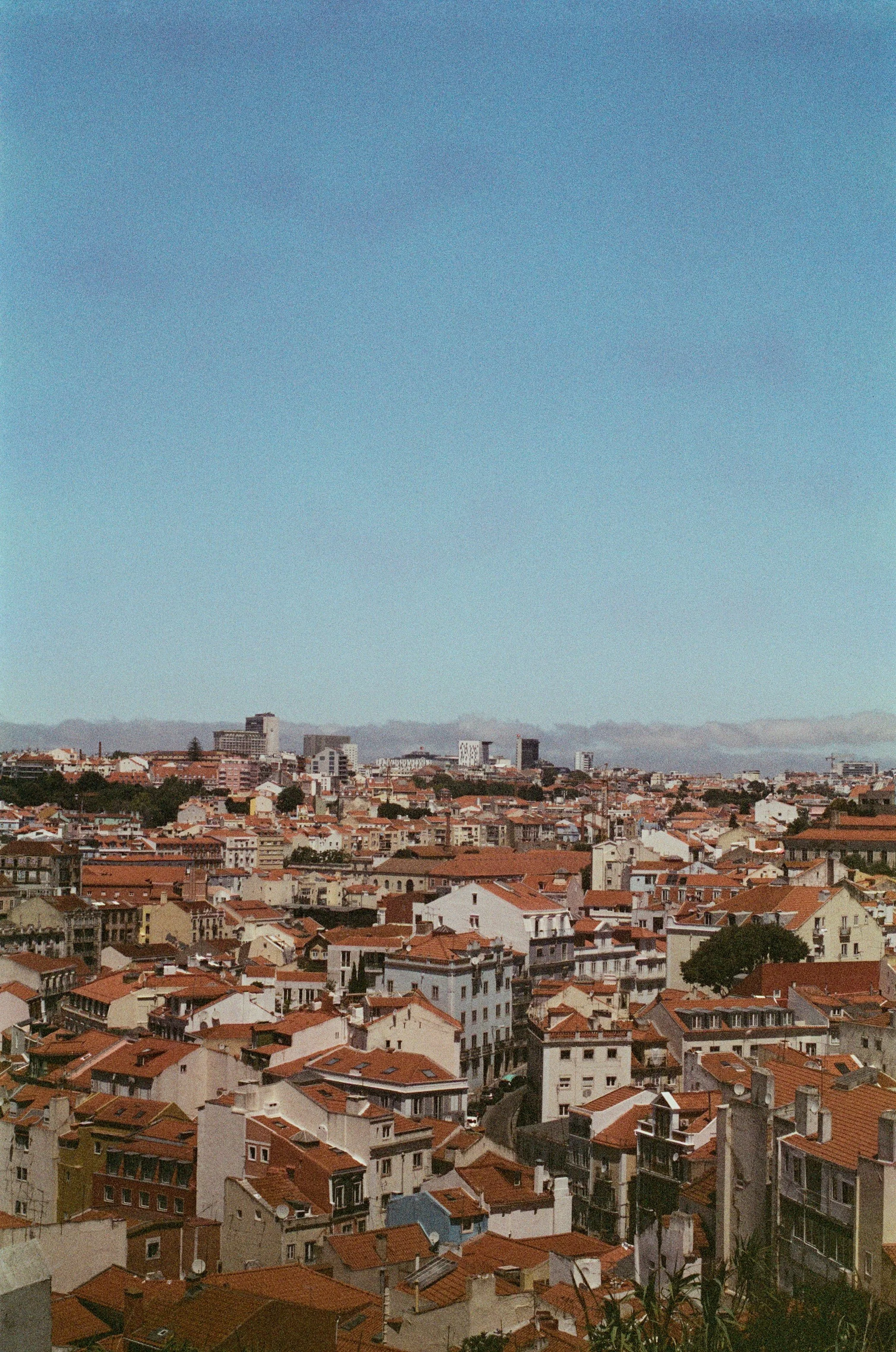 Cityscape with densely packed buildings and red-tiled roofs under a clear blue sky, with some taller buildings and cranes in the distance.