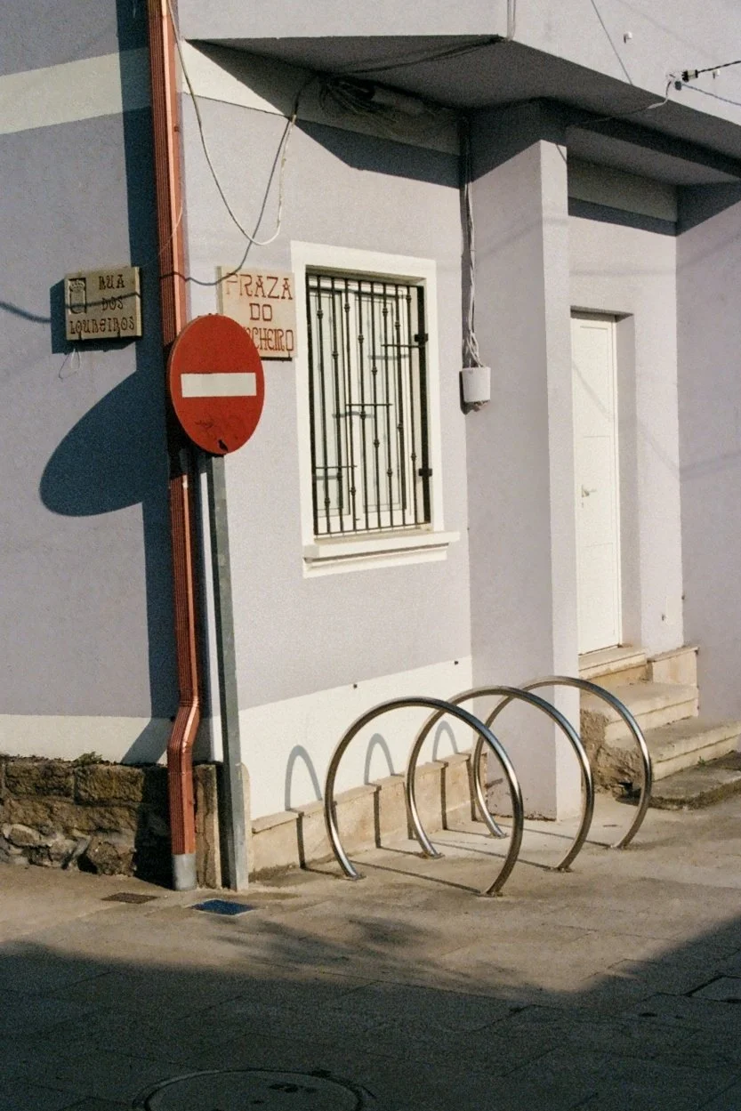 Street corner with a building having a barred window, a white door, and signs. One sign reads "Praza do Cheirido" and another reads "Rua dos Loureiros." There are three metal bike racks in front of the building and a no-entry traffic sign. Shadows ca