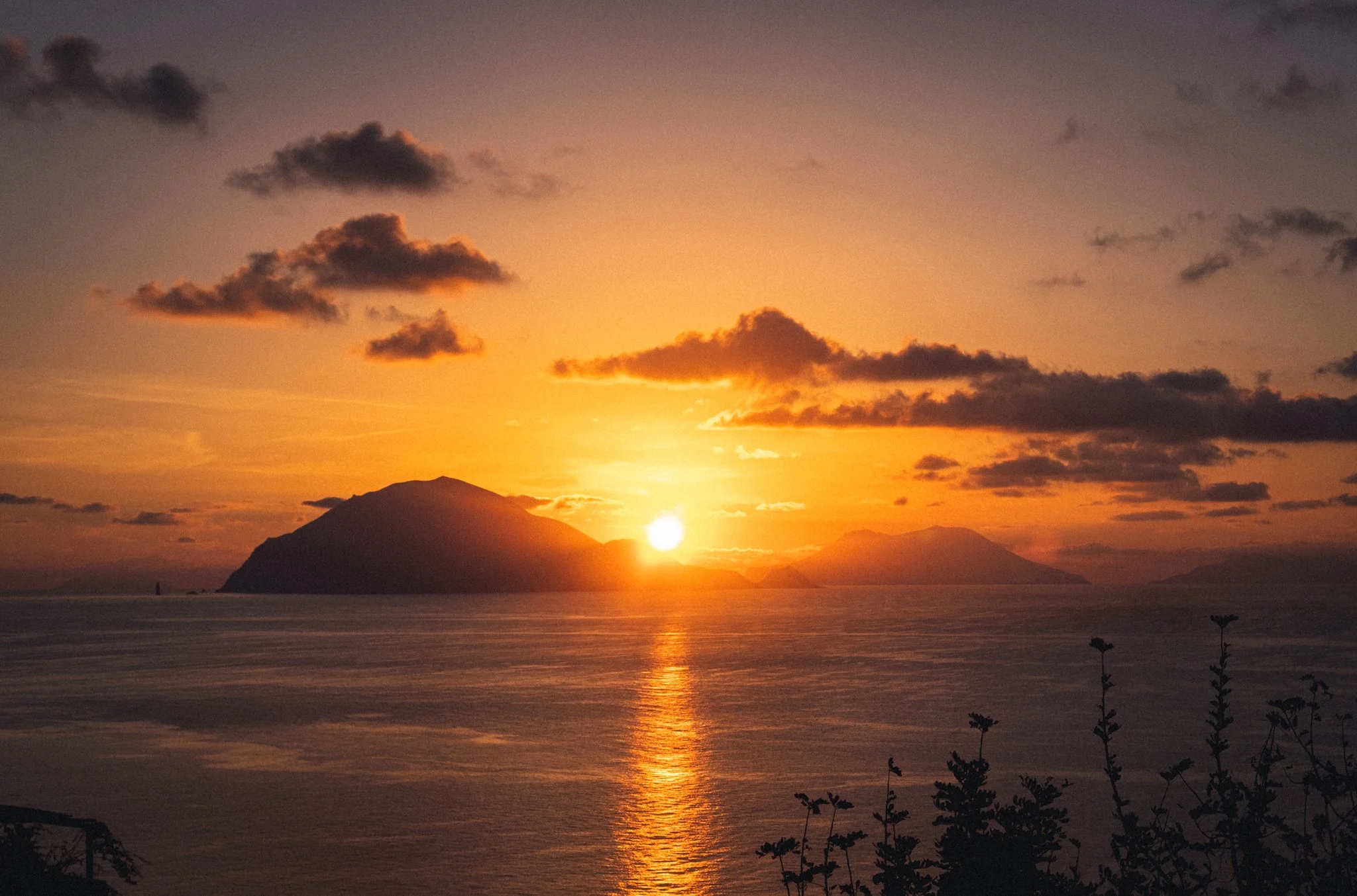 Sunset over a body of water with dark clouds in the sky, hills or mountains silhouette in the background, and some plants or bushes in the foreground.