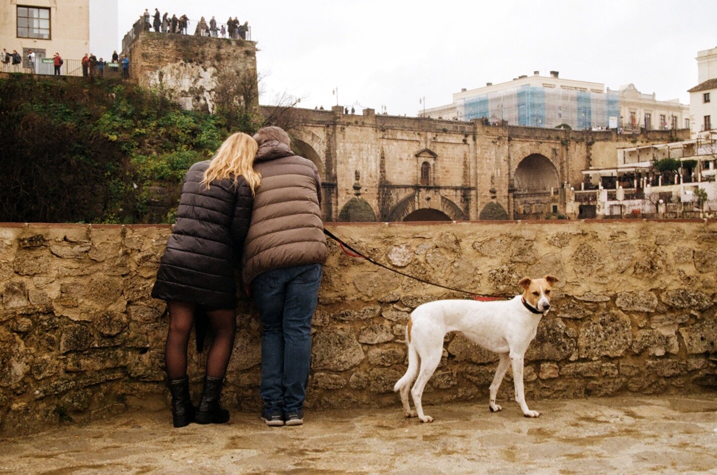Two people, a woman and a man, stand together leaning on a stone wall, looking at an old stone bridge with arches in the background. The woman has blonde hair and is wearing a black puffer jacket with black tights and boots. The man has gray hair and