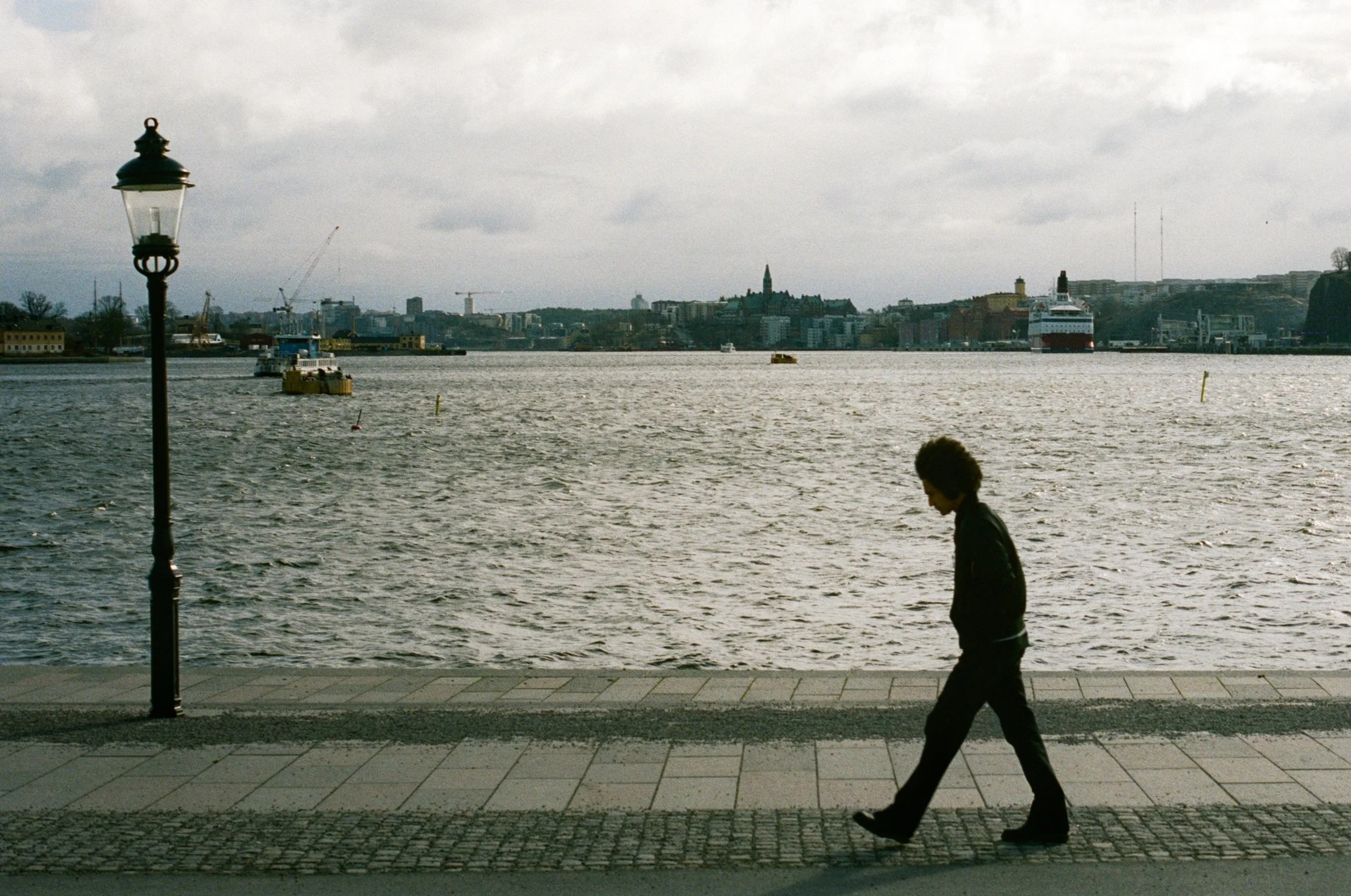 A person walking along a waterfront promenade near a lamp post, with a body of water and city skyline in the background.