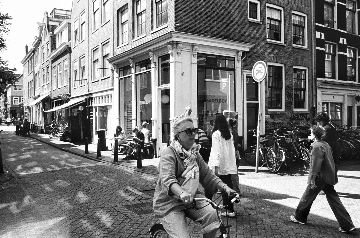 A woman riding a bicycle on a busy city street with pedestrians walking on the sidewalk. Buildings line the street, with bicycles parked along the sidewalk and storefronts visible, including one named 'BAGELBOY'.