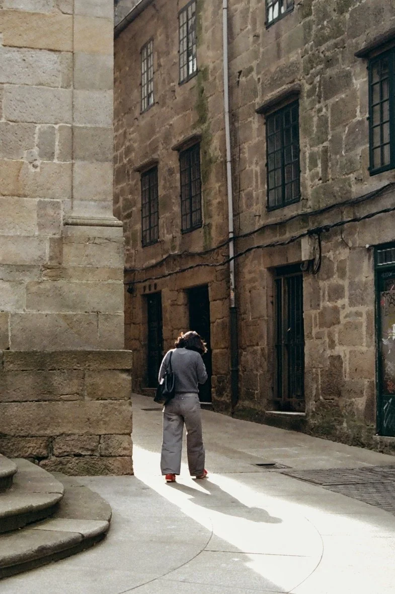 A woman walking down an alleyway between historic stone buildings with barred windows and a drain on the ground.