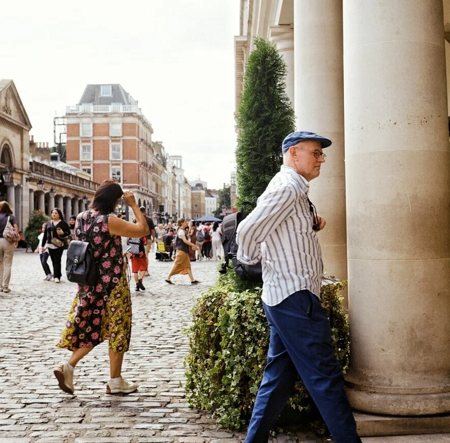 A man leaning against a column on a busy city street, with a woman walking by in the background.
