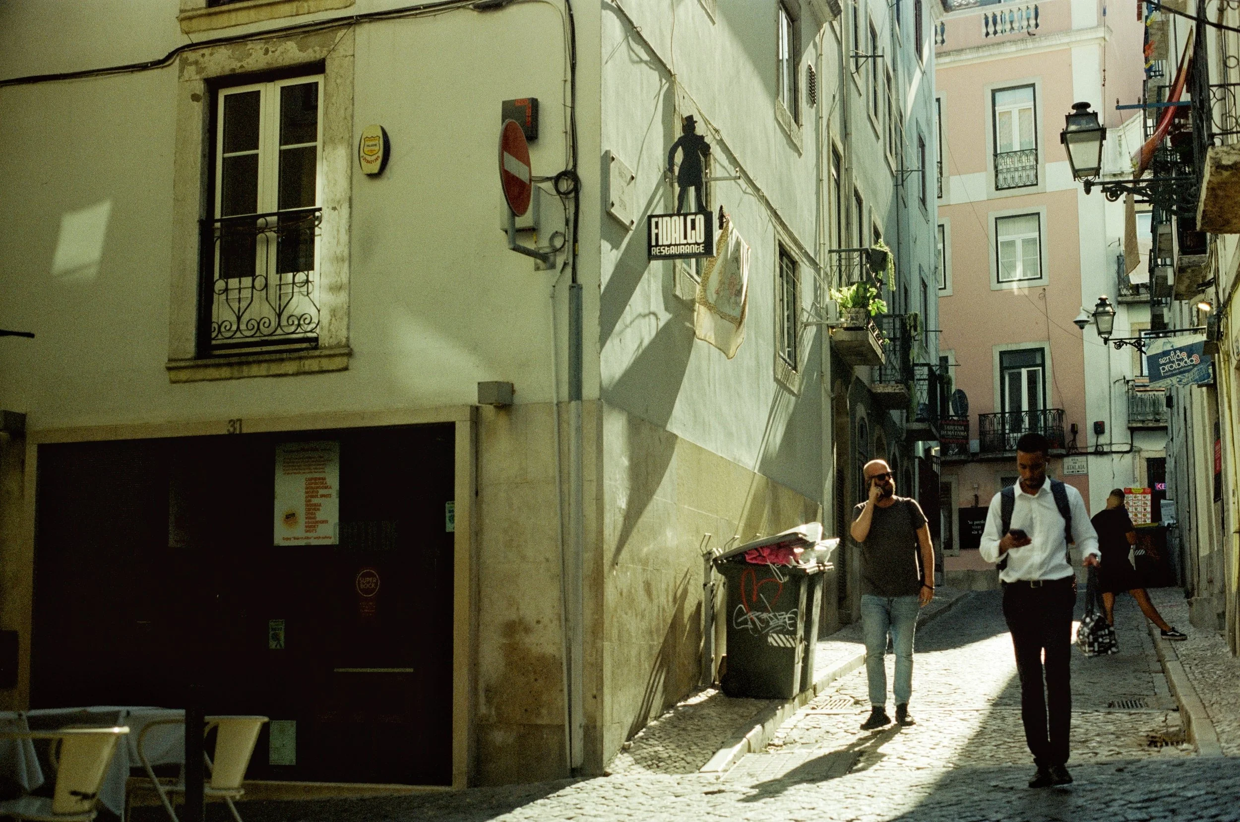Street scene in a European city with cobblestone streets, old buildings with balconies, and a few people walking, including a man talking on the phone and another using a mobile phone.
