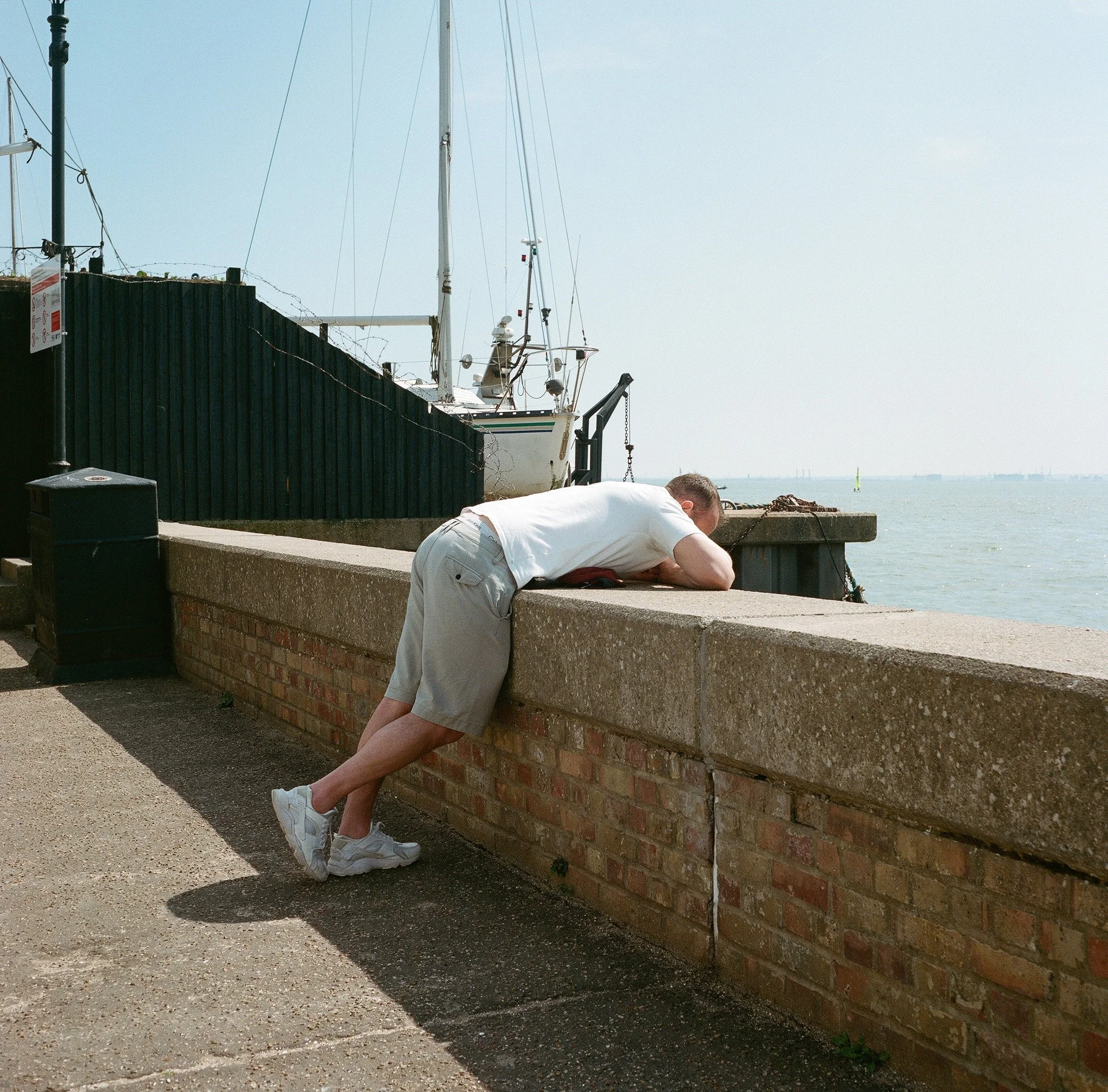 A man leaning over a brick and concrete harbor wall, looking at the water and boats in the distance.