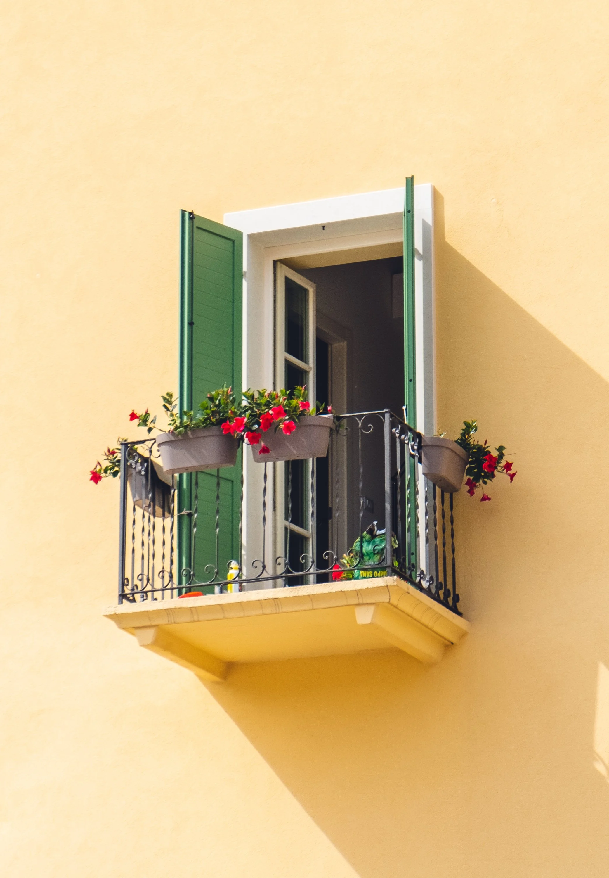 Small balcony with open door and green shutters, decorated with pink and red flowers in grey planters, attached to the yellow building wall.