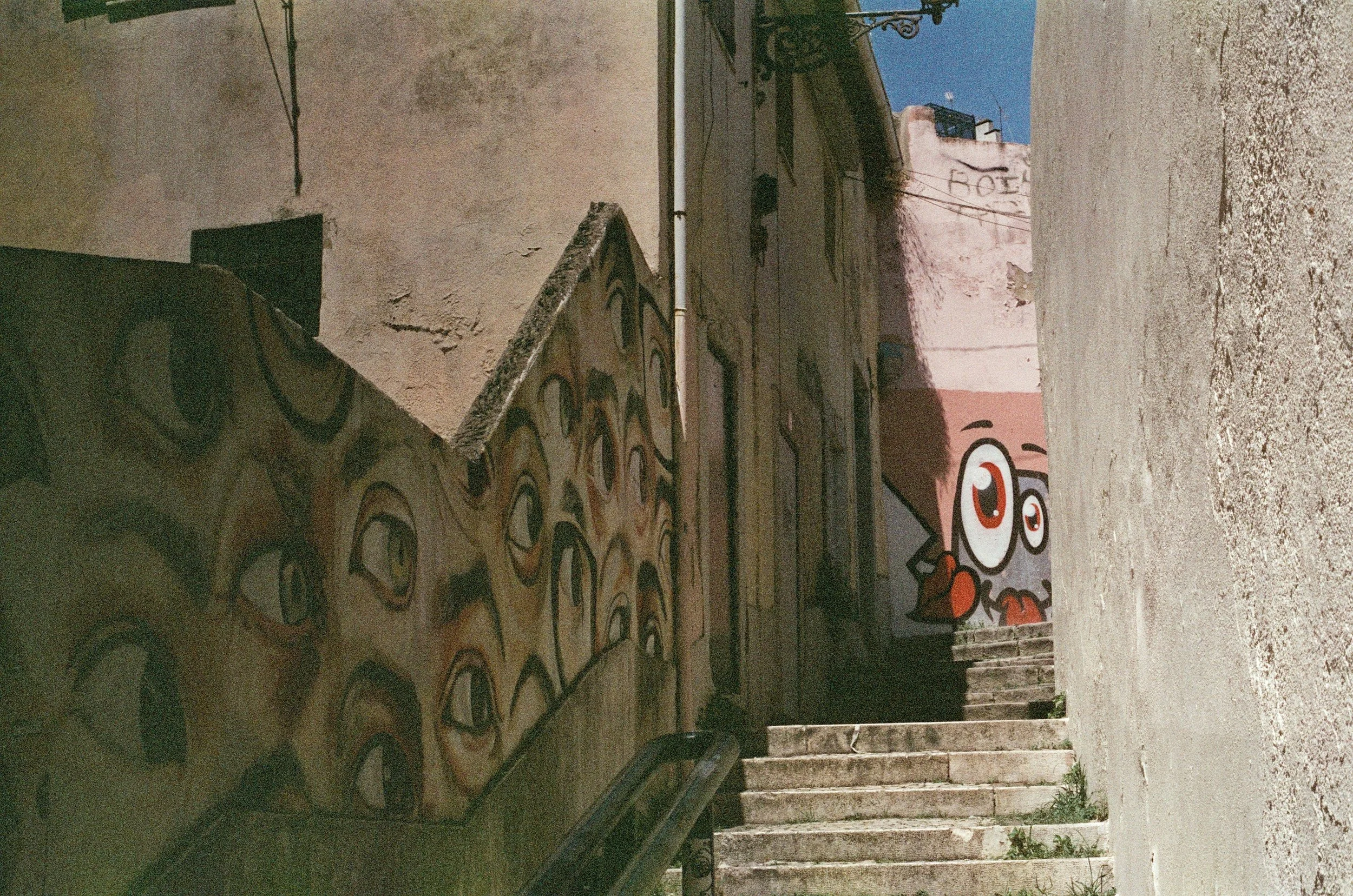 Street alley with graffiti art on walls, one wall features multiple cartoon-like eyes, and a cartoon character with glasses and a brown jacket on the stairs, under a blue sky.