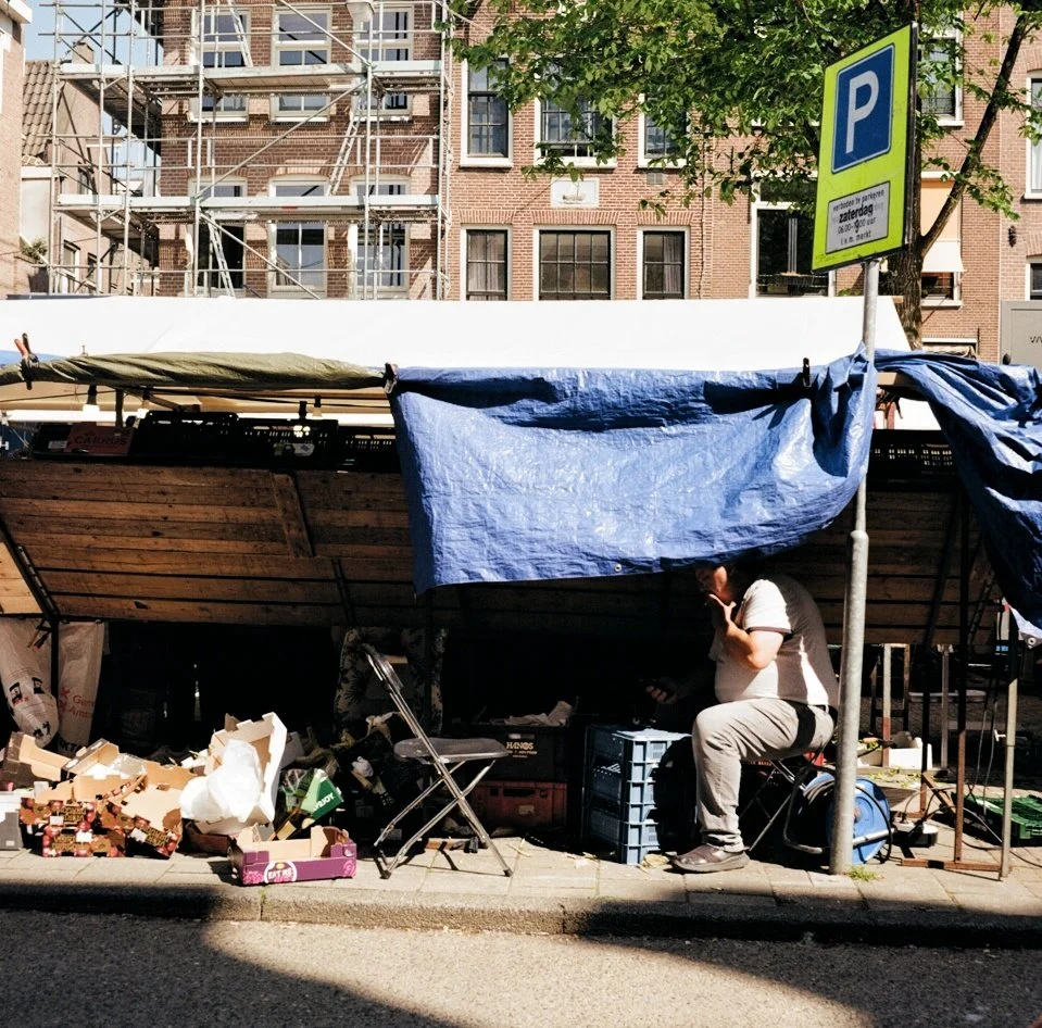 A small street market stall with boxes, crates, and bags, shaded by a blue tarp, with a man sitting and talking on his phone beneath the shade. There is a parking sign and high-rise apartment buildings in the background.