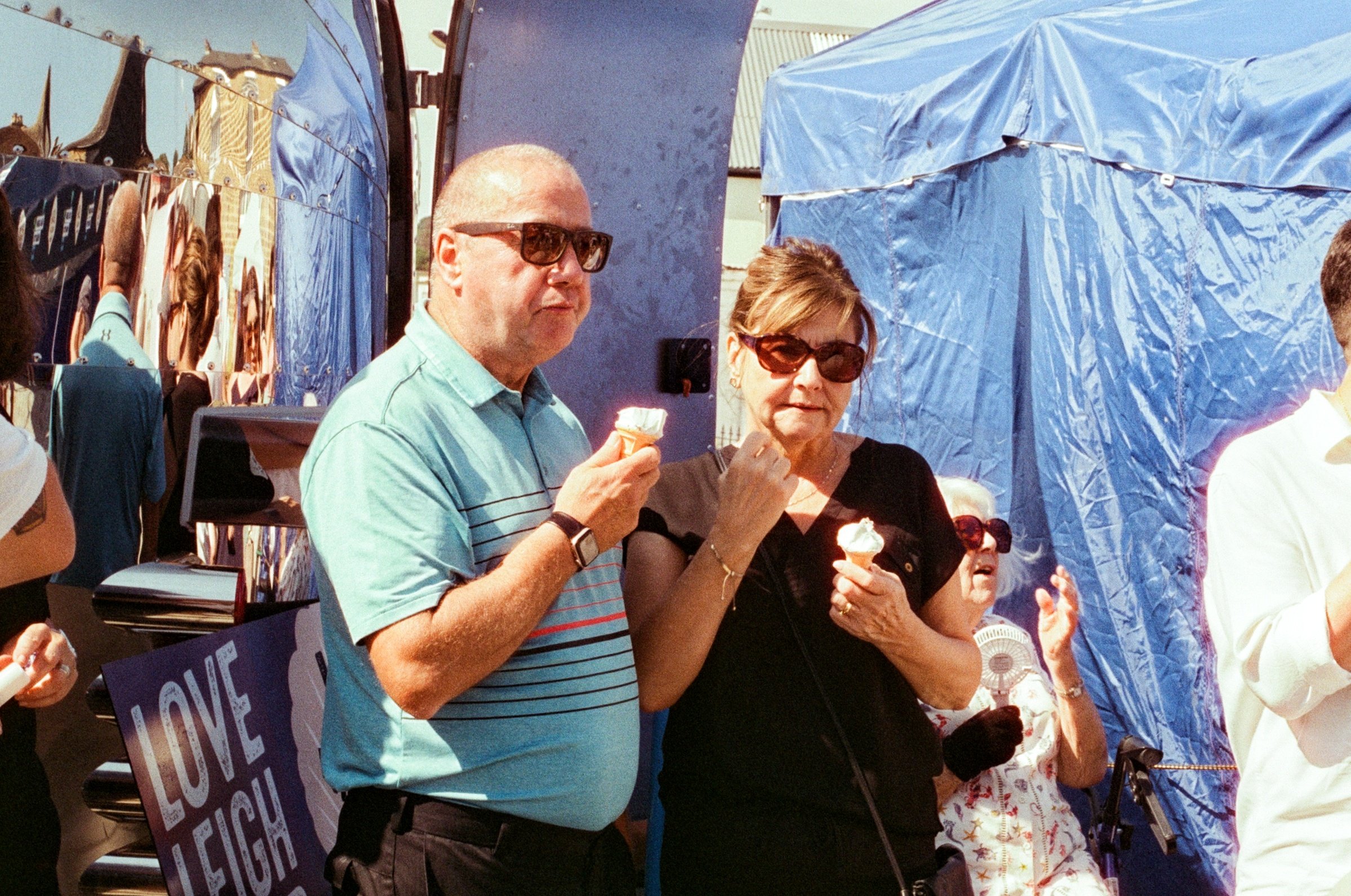 People at an outdoor event eating ice cream, with a blue tent and reflective surfaces in the background.