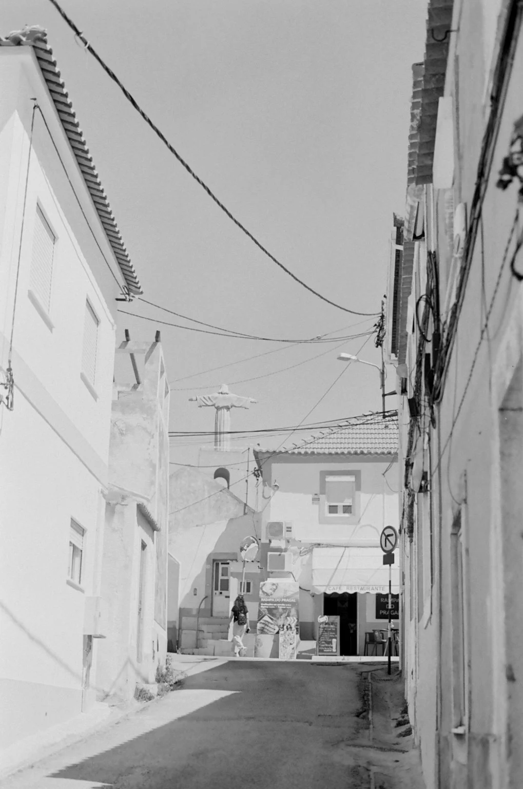 A narrow street in a small town, with buildings on both sides, a person walking, and a statue of Christ with open arms in the background.