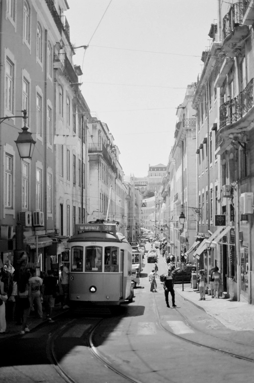 Black and white photo of a busy city street with a tram in the center, surrounded by pedestrians, parked cars, and tall buildings with balconies.