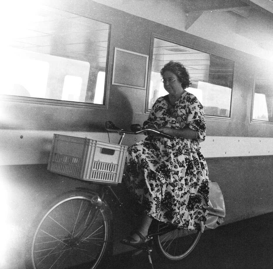 A woman in a floral dress riding a bicycle with a basket in front, parked in front of a train.