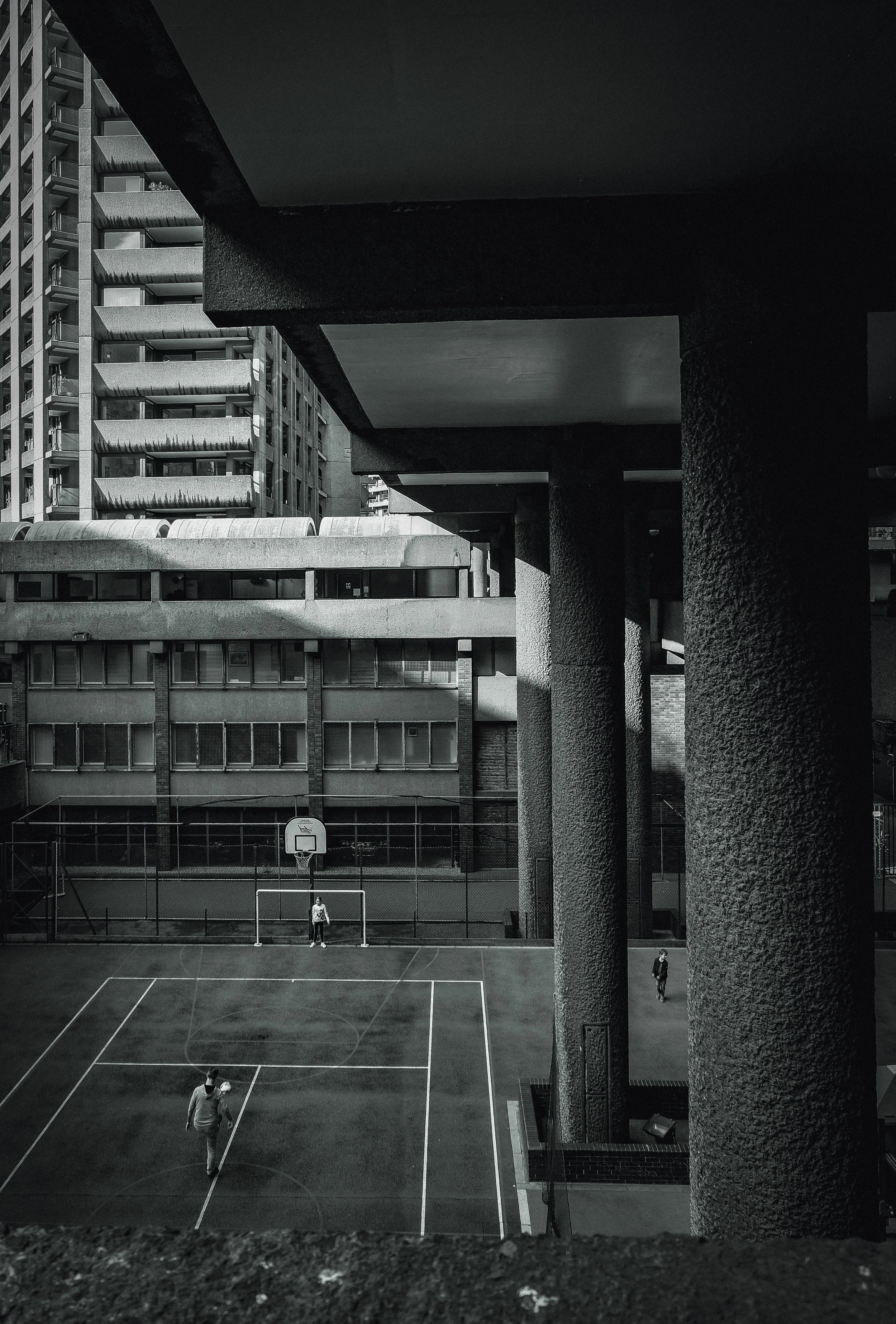 Black and white photo of a basketball court between tall apartment buildings, with three people on the court and a basketball hoop.