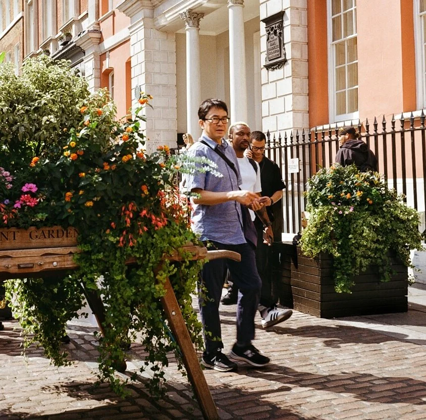 Three people walking past flower planters on a cobblestone street in front of a building with columns and pink walls.