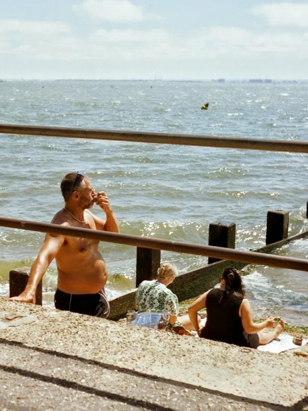 People relaxing by the water at a beach or pier, with one man laying back and another person sitting on the sand and an older woman sitting by the water.