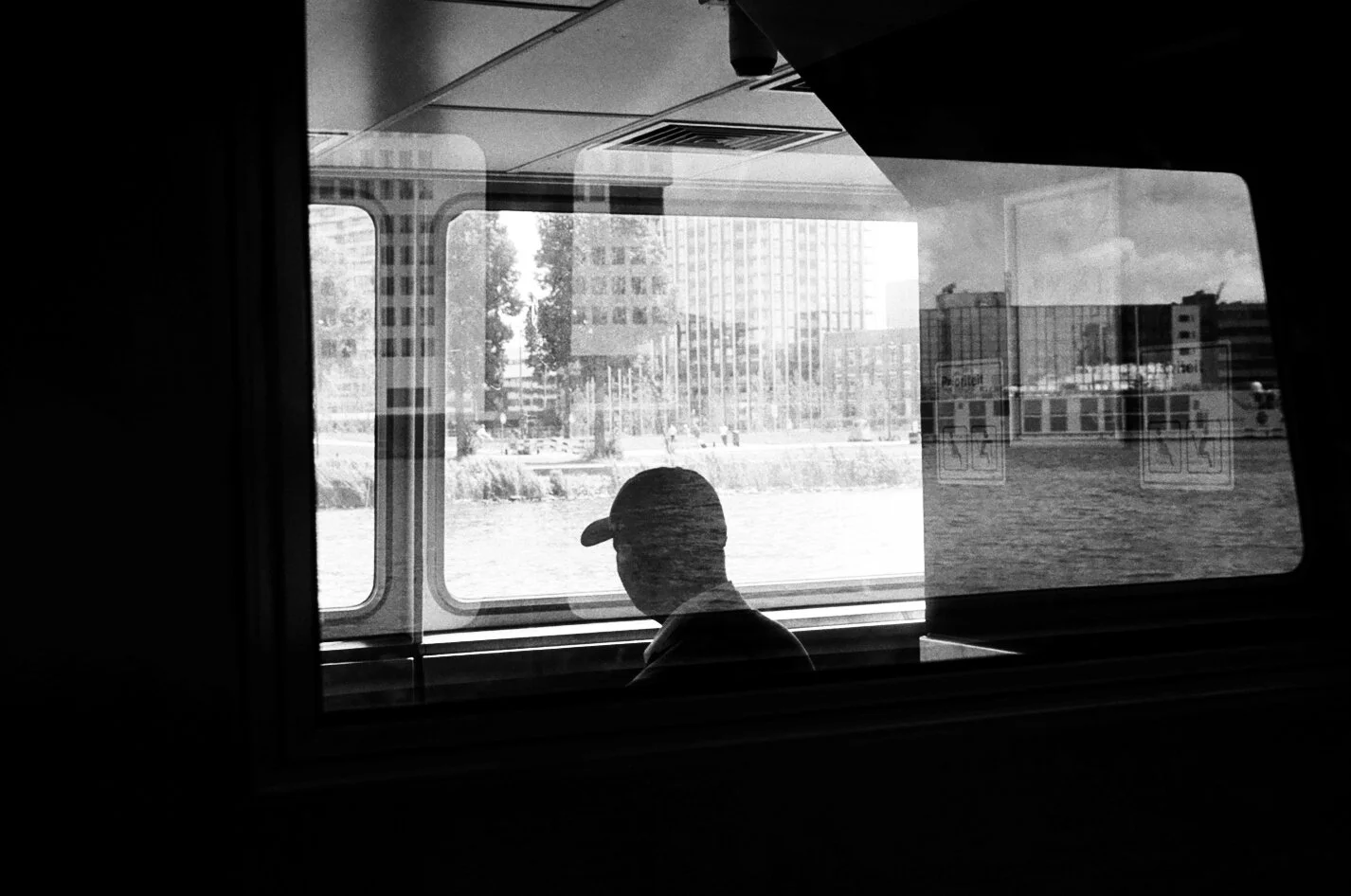 Black and white photo of a man wearing a cap sitting by a window on a boat or train, with city buildings and water outside.