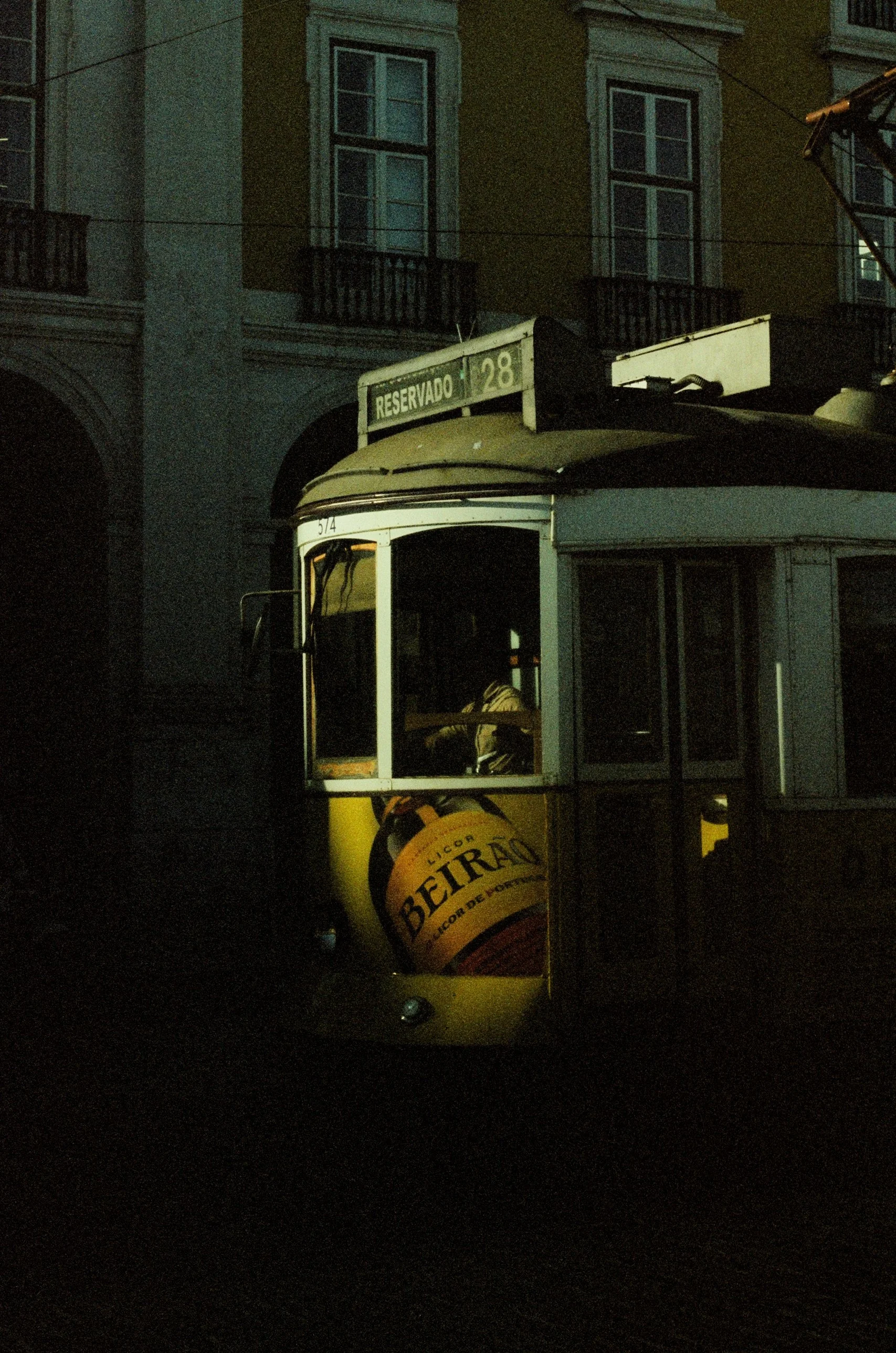 Nighttime scene of a vintage yellow tram with a sign reading 'Reservado' and a beer advertisement on the front, with a dark urban background of buildings with windows.