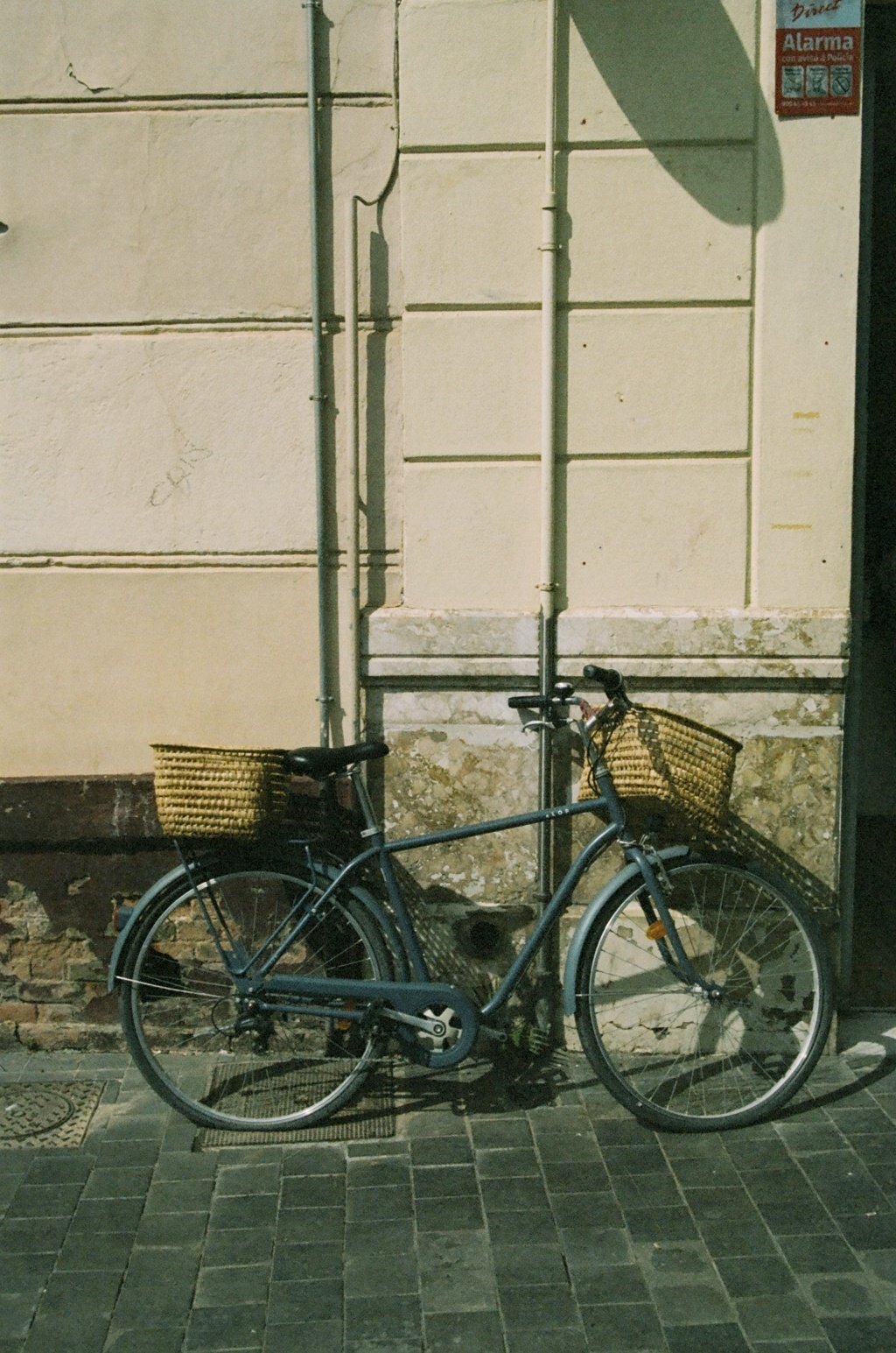 A bicycle with wicker basket baskets parked against a wall on a sidewalk at night.