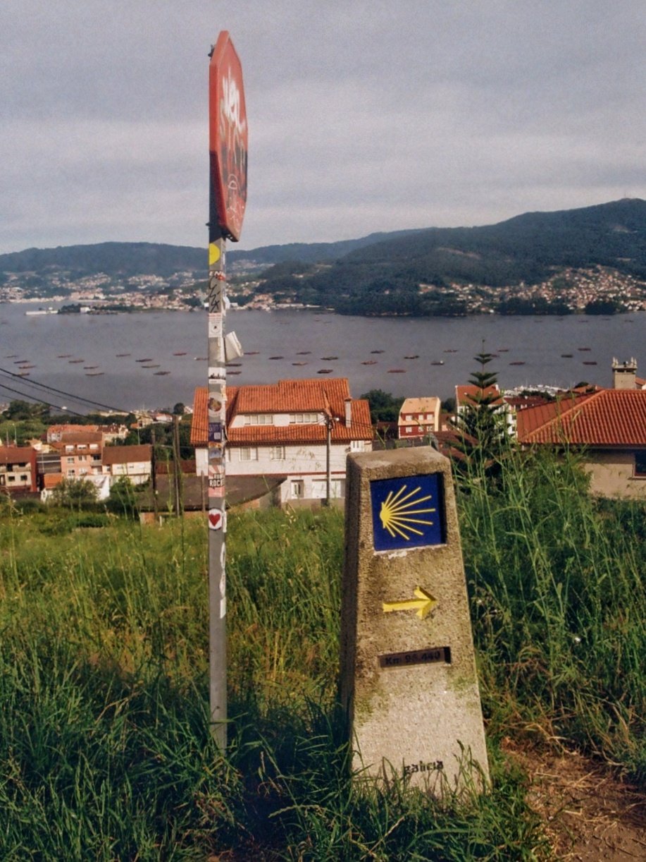 A stone marker with the yellow scallop shell symbol and an arrow, indicating the Camino de Santiago trail, set on grassy terrain with houses and a lake in the background.
