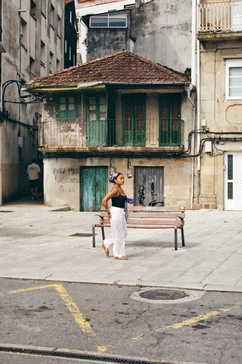 A woman in black top and white pants walking in front of a park bench on a sidewalk in an urban area with old buildings, graffiti, and graffiti on the wall behind her.