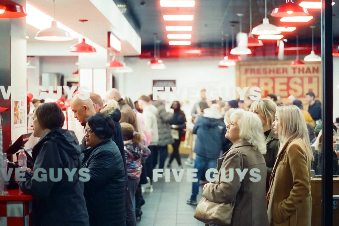 People standing in line inside a restaurant or fast-food place, with red and white decor and the sign 'FRESHER THAN FRESH' visible in the background.