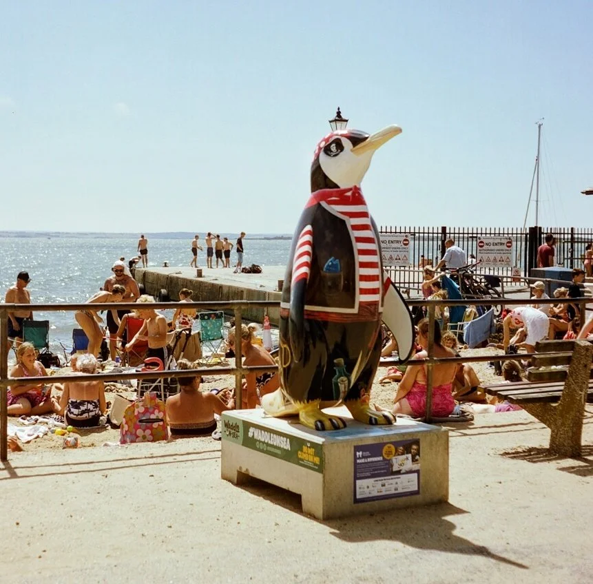 A penguin statue dressed in a red and white striped scarf and jacket, standing on a pedestal at a beach with people sunbathing, swimming, and relaxing near the water and a pier with boats and railings.