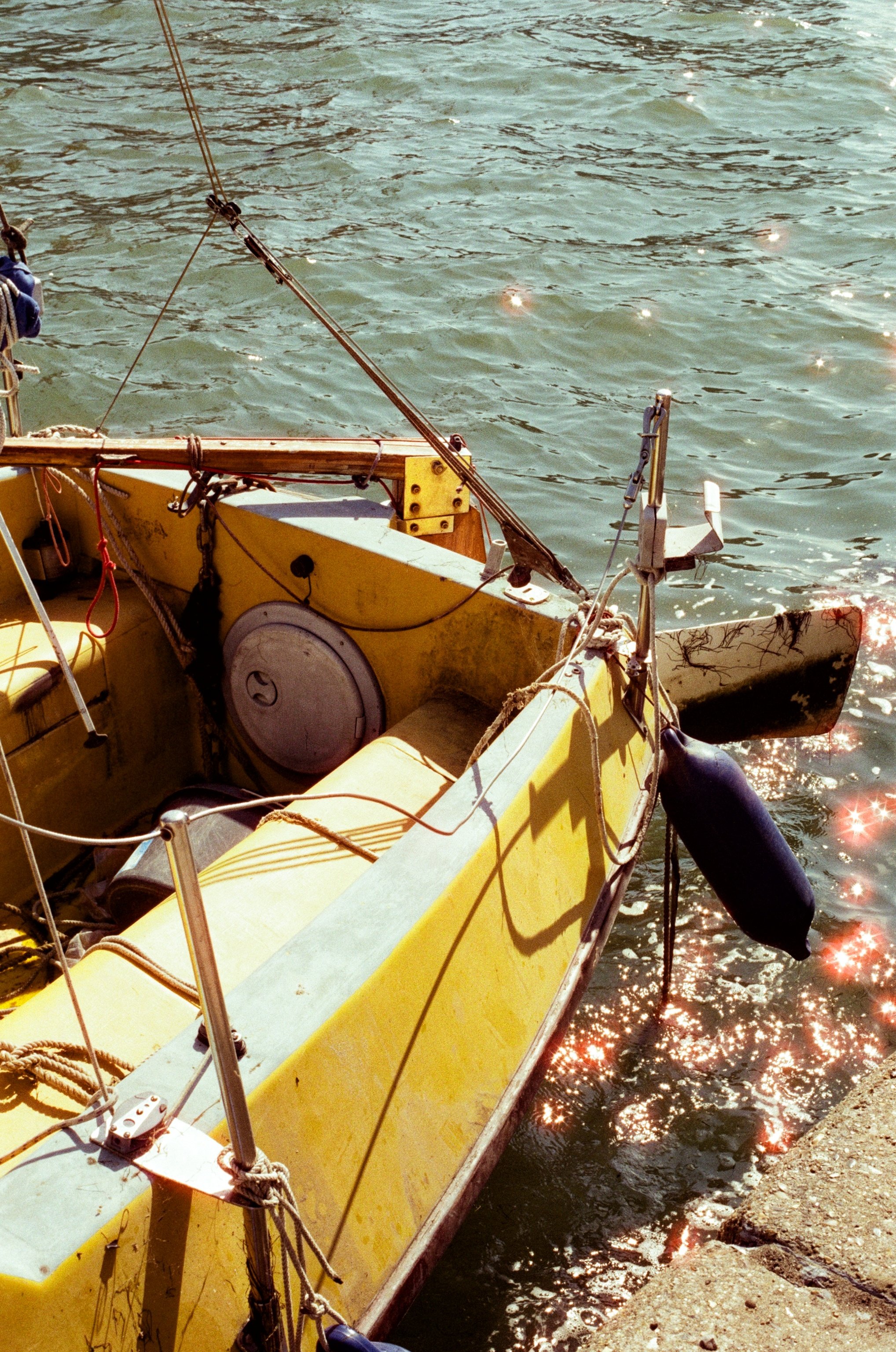 A yellow sailboat docked next to a concrete pier on a sunny day, with the water reflecting sunlight and sparkles visible around the boat.