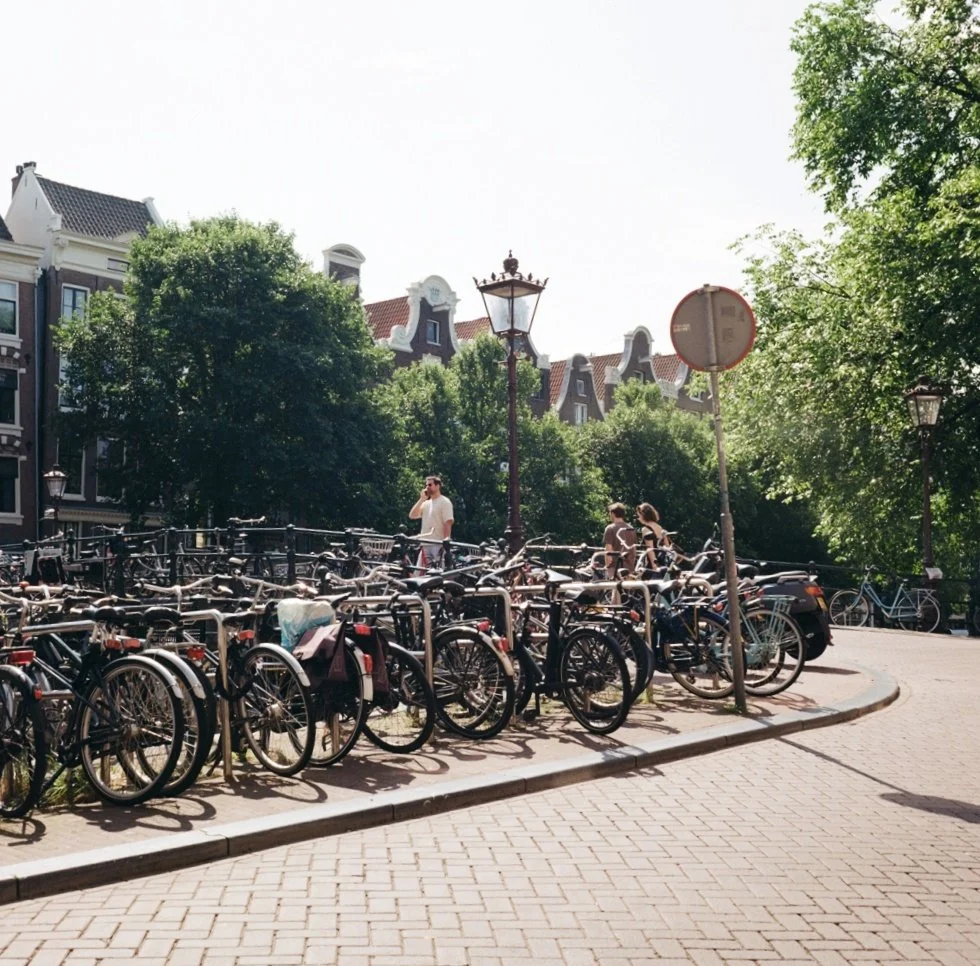 A street scene with multiple parked bicycles, a few people walking and talking, and historic European-style buildings in the background under a bright sky.