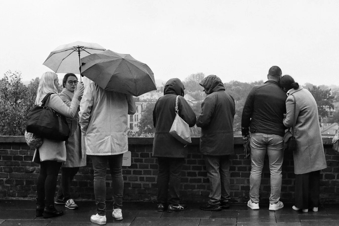 People standing on a bridge, viewing the cityscape through a brick barrier on a rainy day. Some are holding umbrellas, and they are dressed in coats and jackets.
