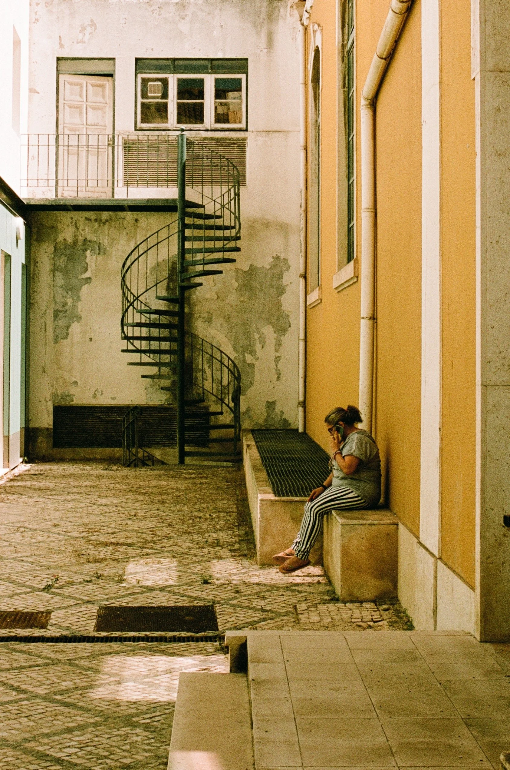 A woman sits on a bench, talking on her cellphone, in an outdoor alleyway illuminated by streetlights. The surrounding walls are a mix of yellow and weathered concrete, with a black spiral staircase leading to an upper level. The ground is paved with