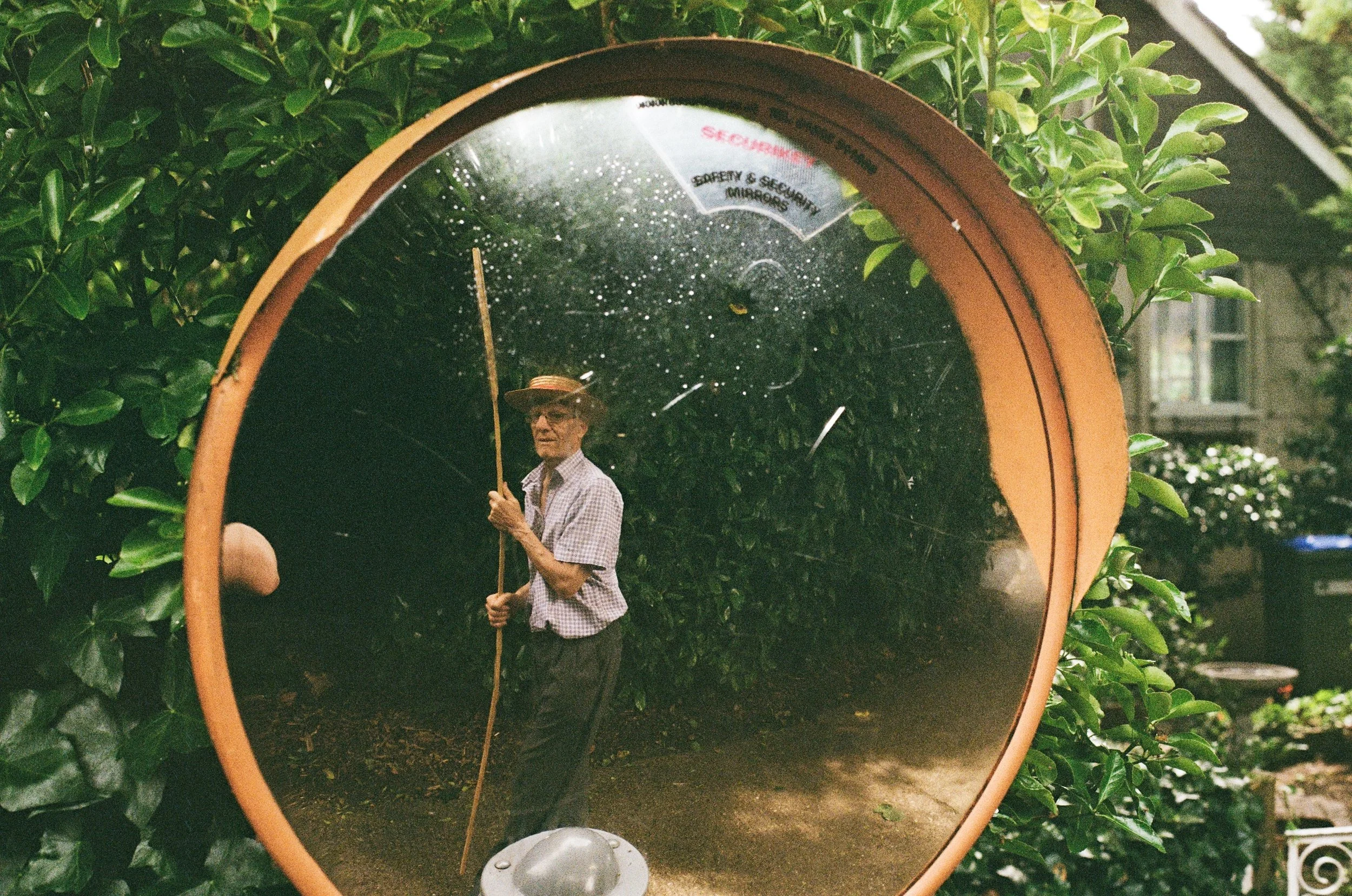 A man wearing a hat and glasses holds a walking stick inside a reflective, curved surface, creating a distorted mirror image of the surrounding greenery and his figure.
