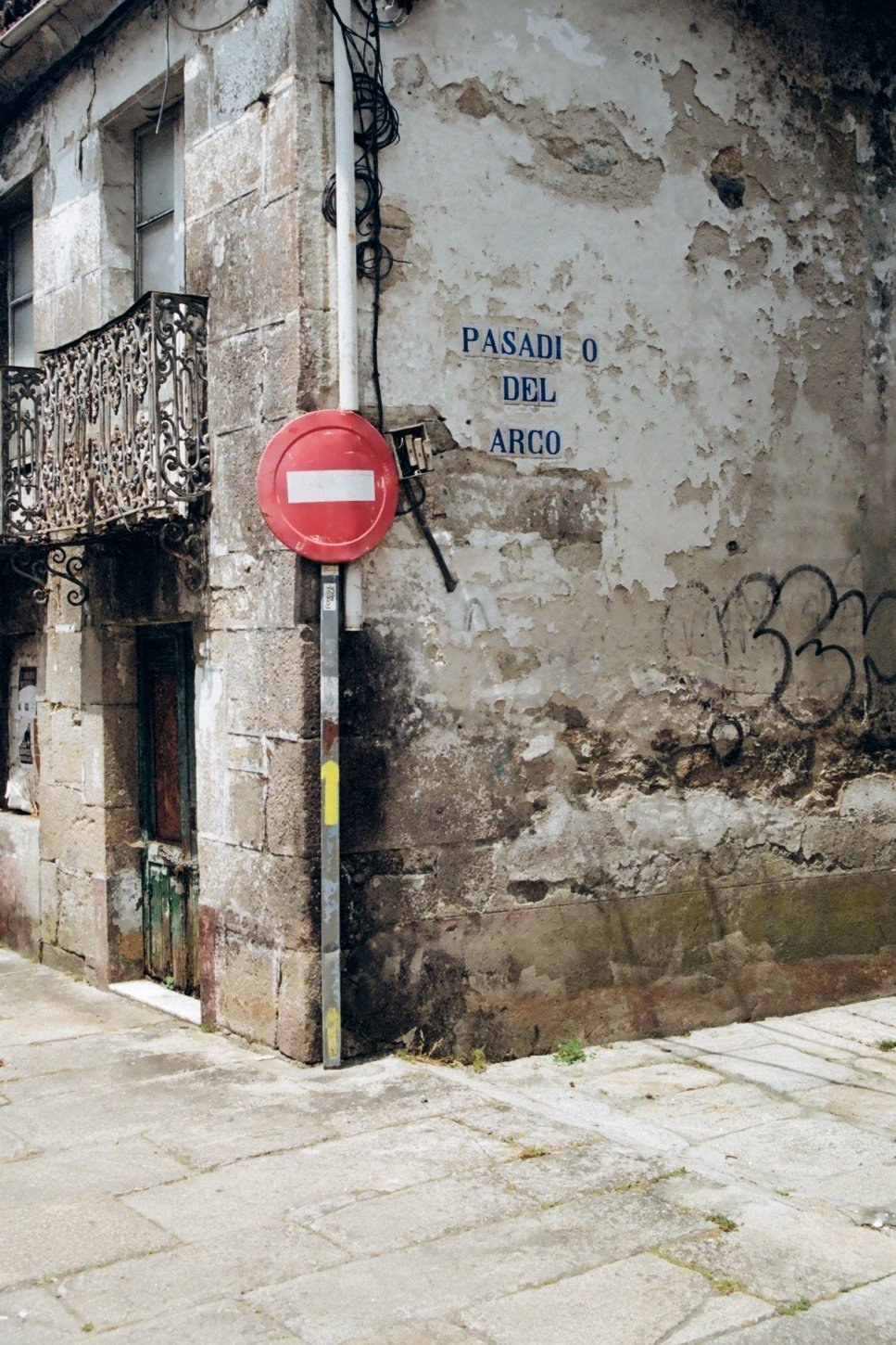A weathered corner building with peeling paint, a small green door, and a black wrought iron balcony. A red 'no entry' sign is mounted on a pole next to the building. The street sign on the wall reads 'Pasad O del Arco'.