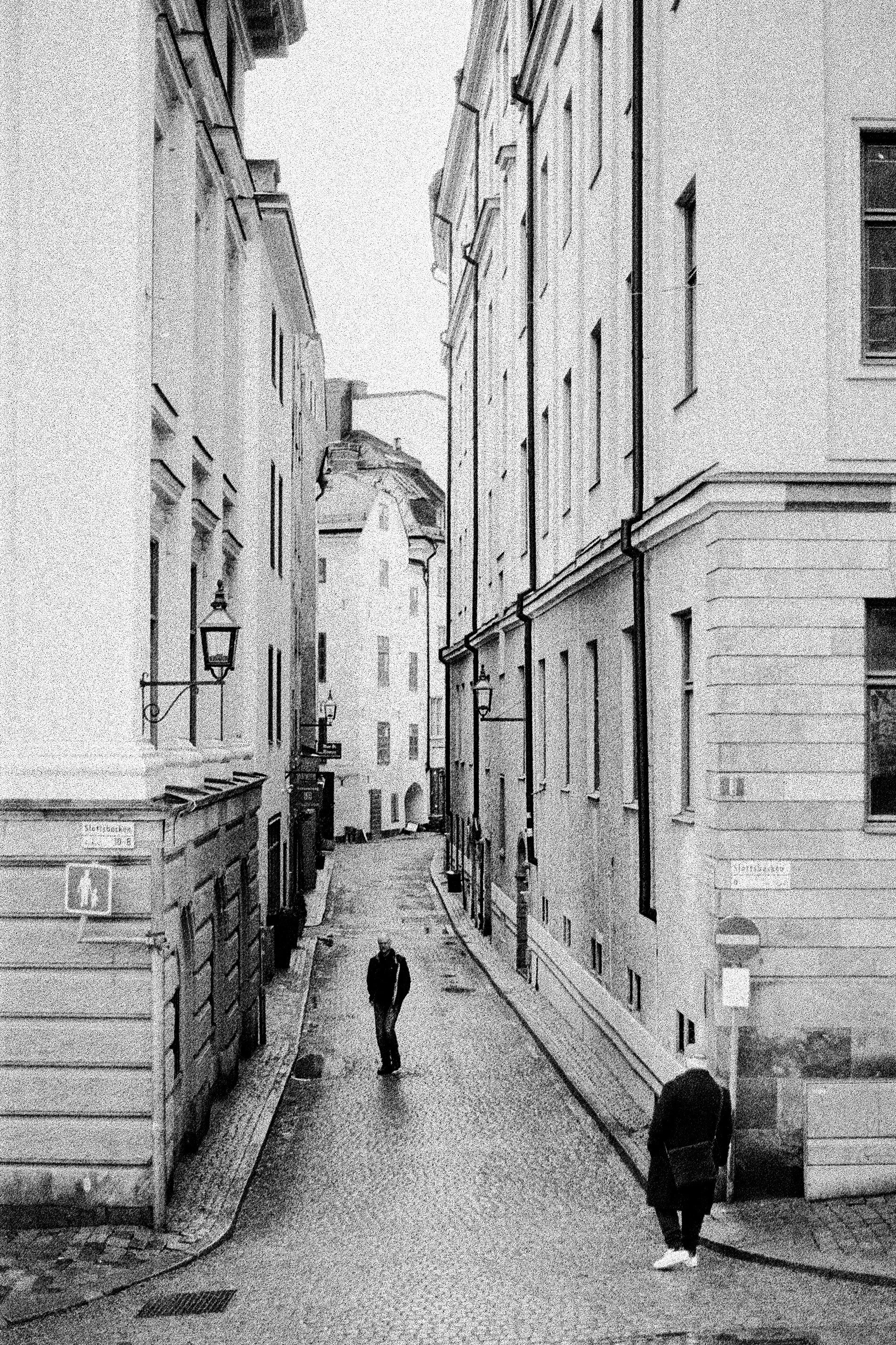 Black and white photo of a narrow city street with two people walking, surrounded by tall old buildings with windows and street lamps.