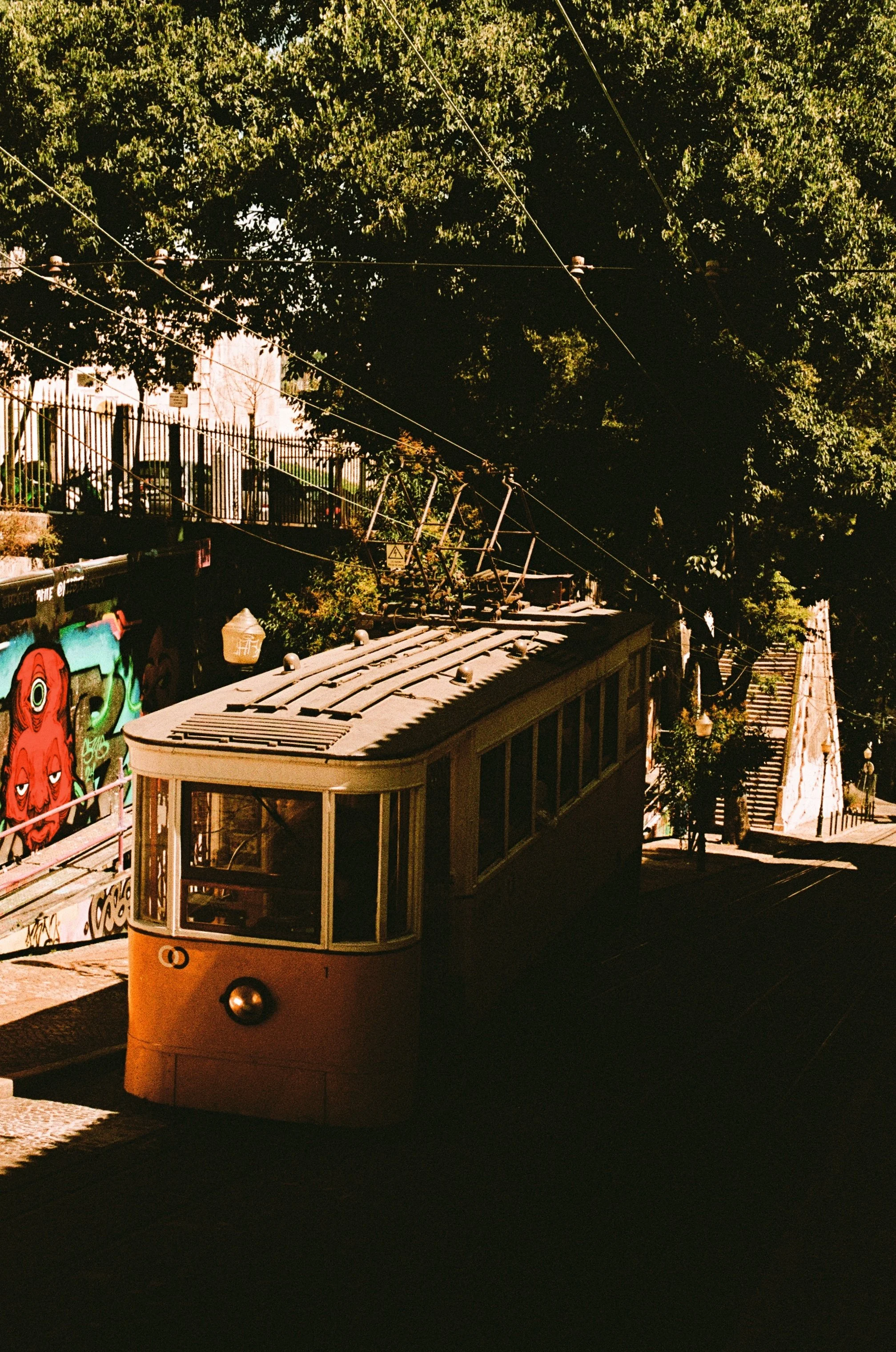 A vintage yellow streetcar traveling on tracks through a city neighborhood under large green trees with stairs and street lamps nearby.