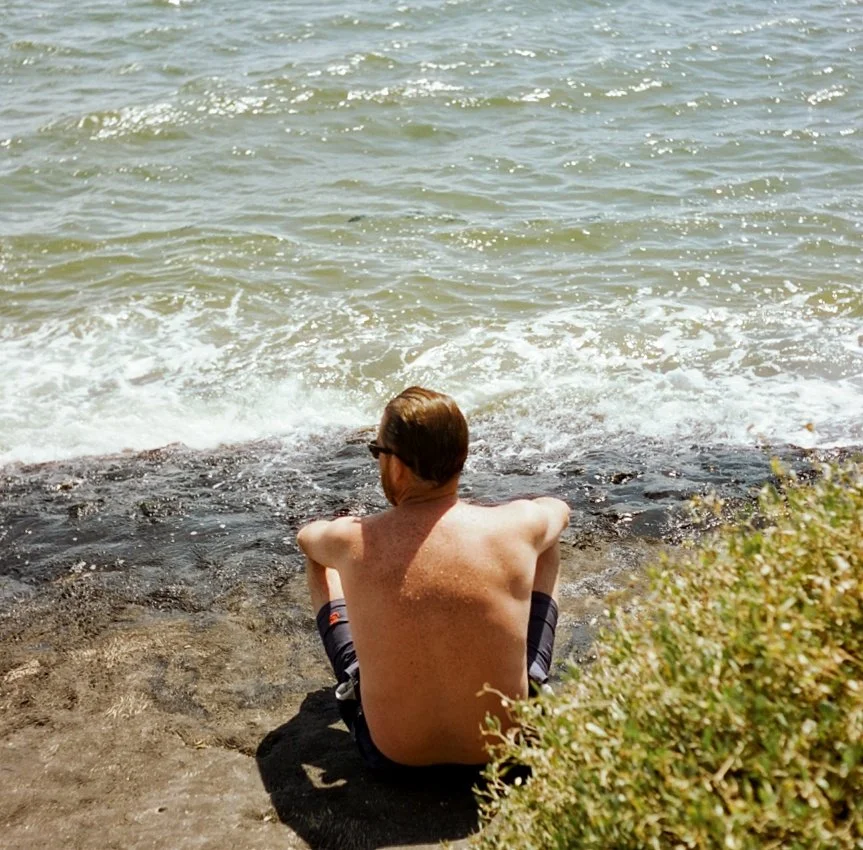 A shirtless man with sunglasses sitting on a rock by the water at the beach, facing away from the camera.