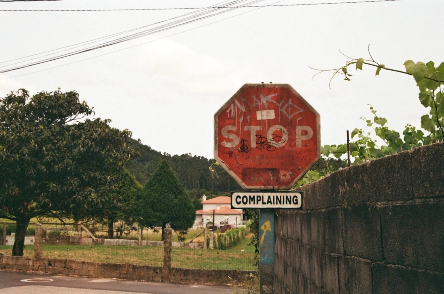 An old rusted stop sign with graffiti and stickers, mounted on a pole next to a brick wall, with a small rectangular sign below reading "COMPLAINING". The background shows trees, a house, and a cloudy sky.